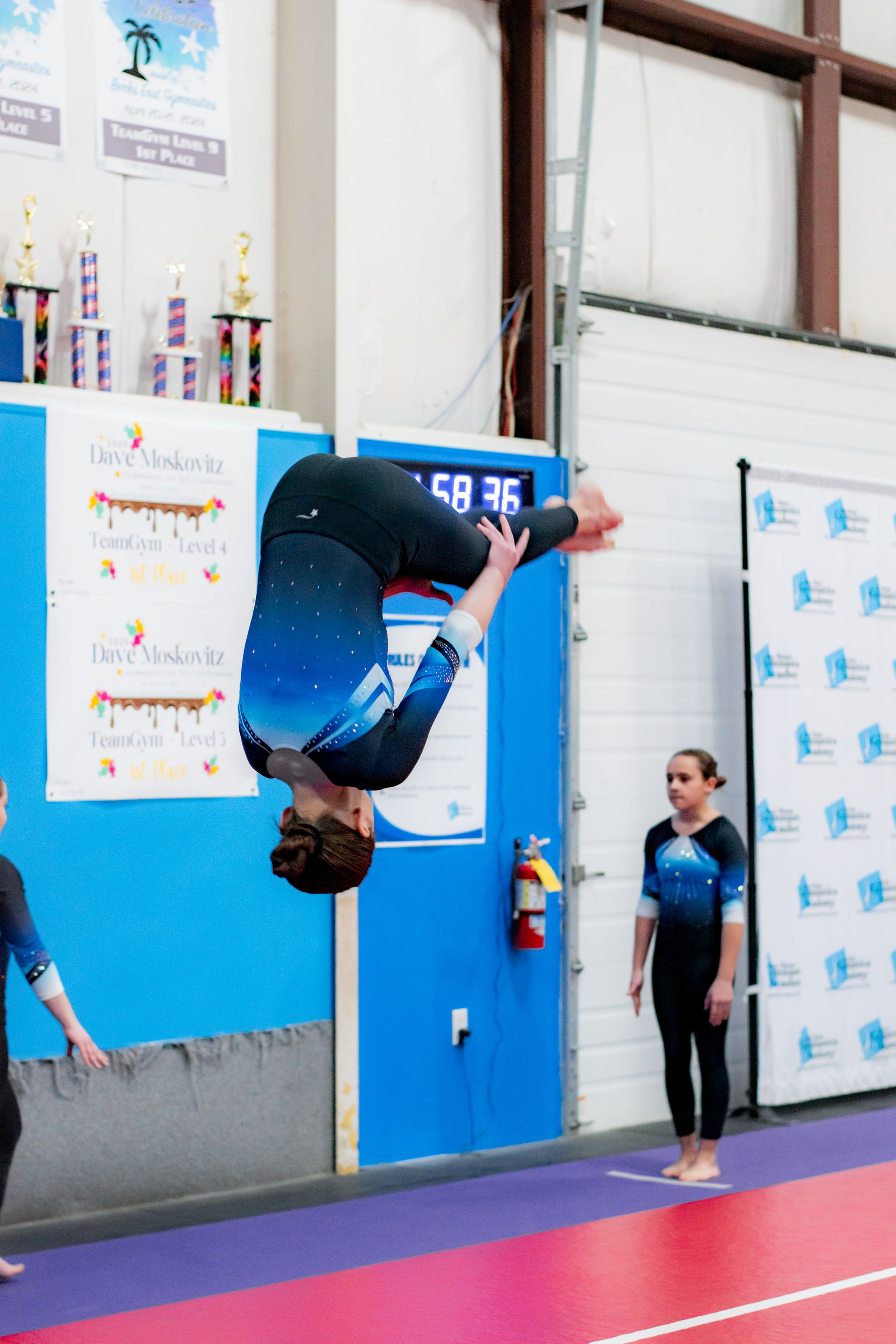 Gymnast mid-flip against a blue wall, with a coach watching nearby.