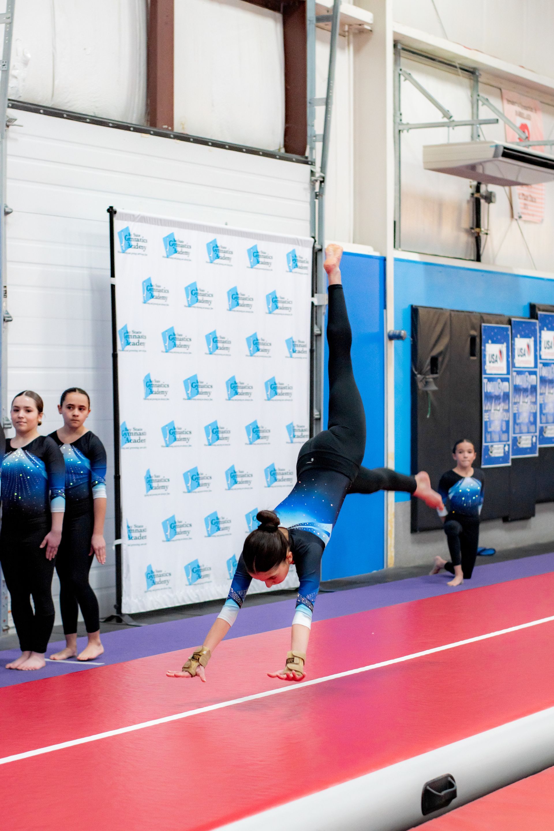 Gymnast doing a handstand on a red mat in an indoor gym while others watch nearby