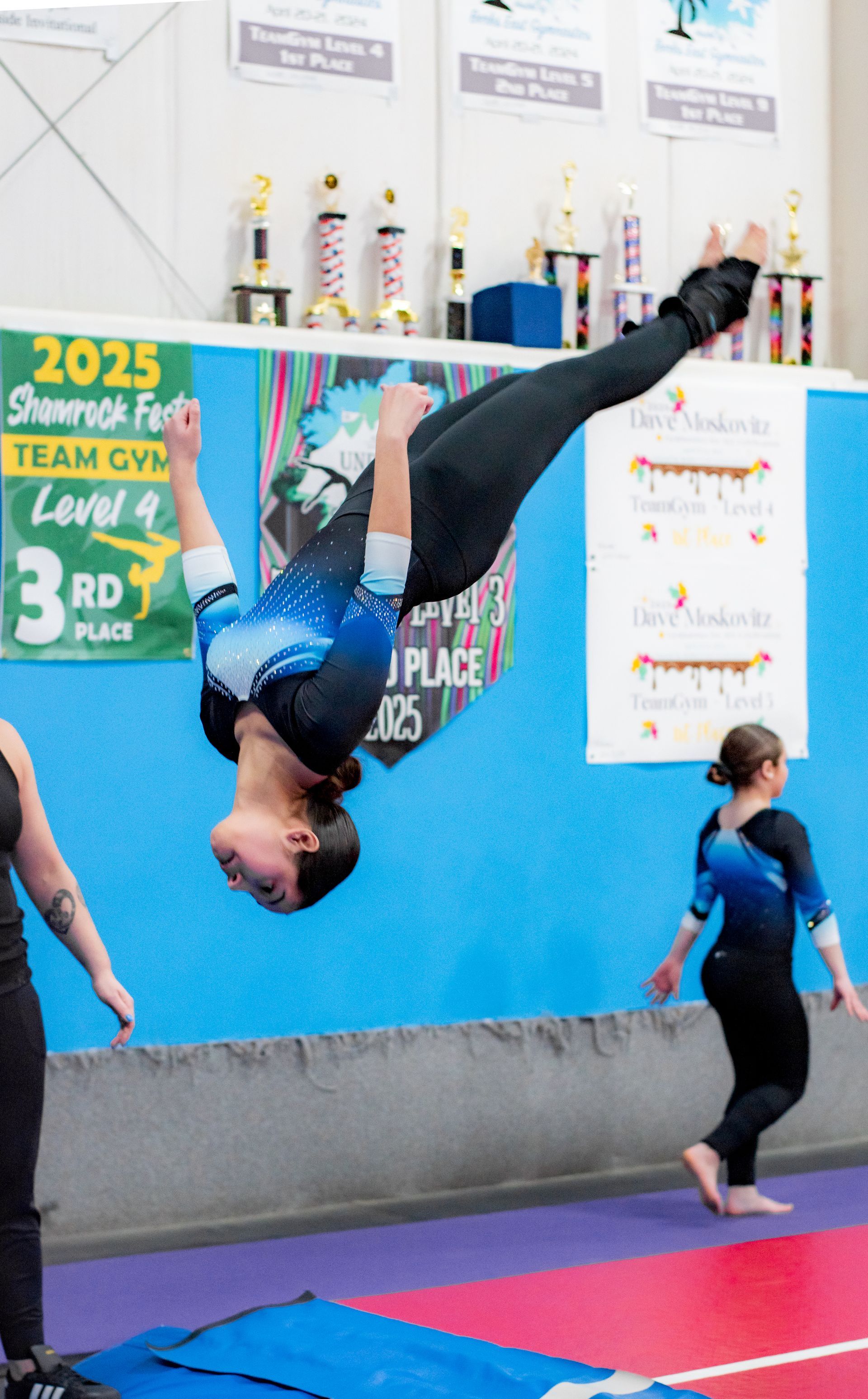 Gymnast mid-backflip in a blue gym, with a child watching nearby and competition banners in the background