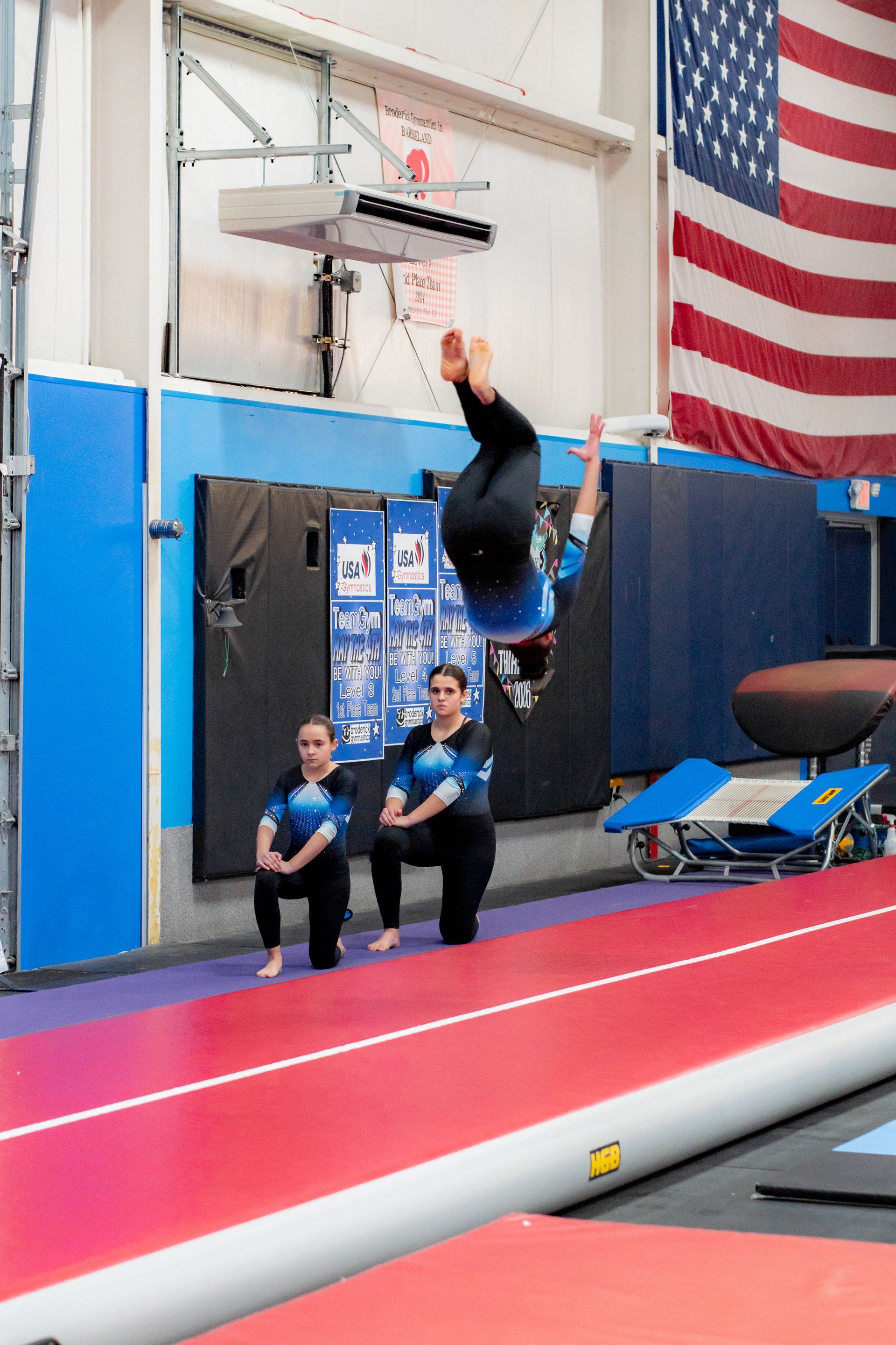 Gymnast swinging on uneven bars in an indoor gym, with coaches watching below.