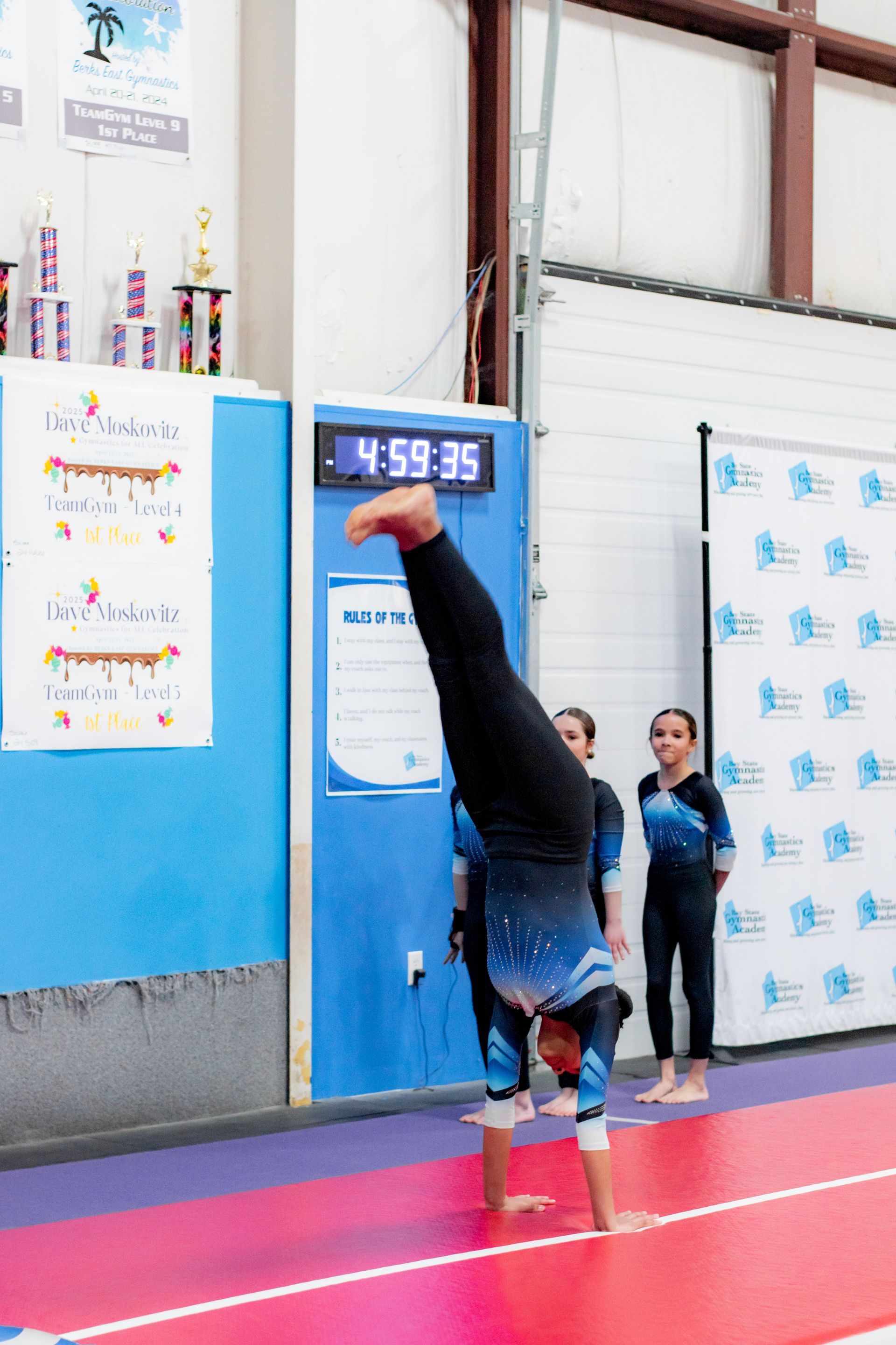 Gymnast doing a handstand on a pink mat indoors, with a scoreboard in the background.