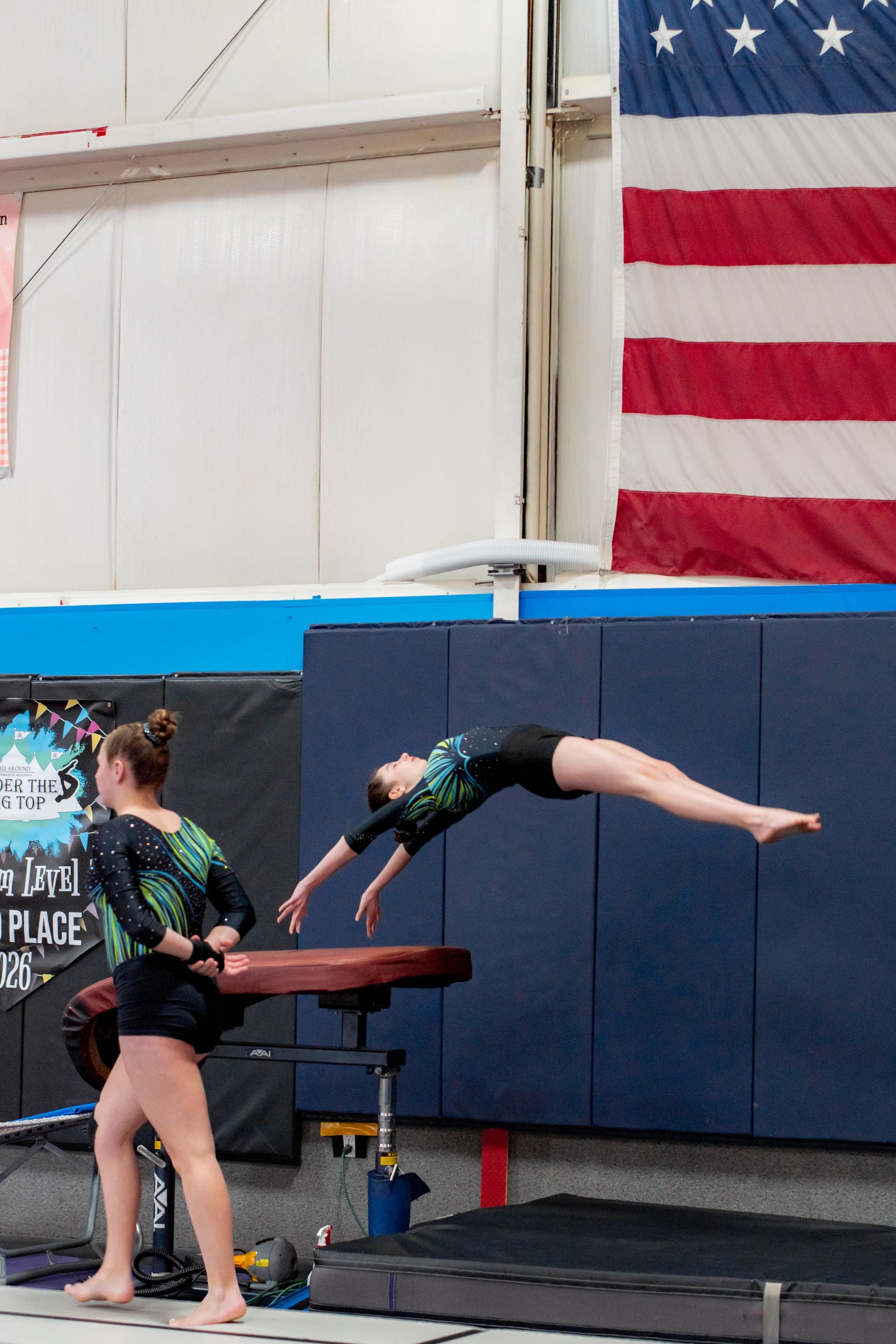 Gymnast mid-backbend over a vault in an indoor gym, with a U.S. flag on the wall.