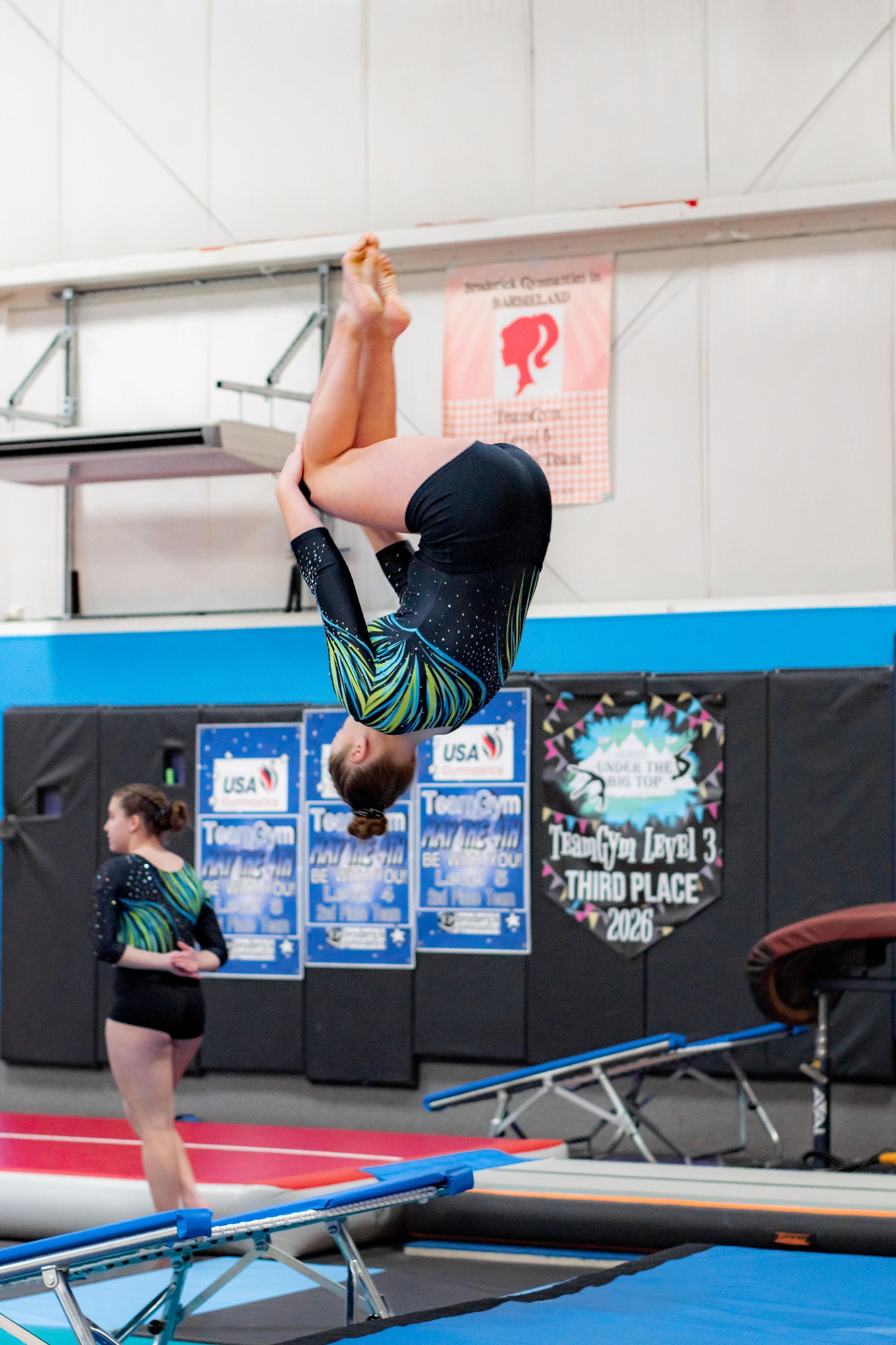 Gymnast in mid-air on a trampoline in an indoor training gym, with another athlete nearby.