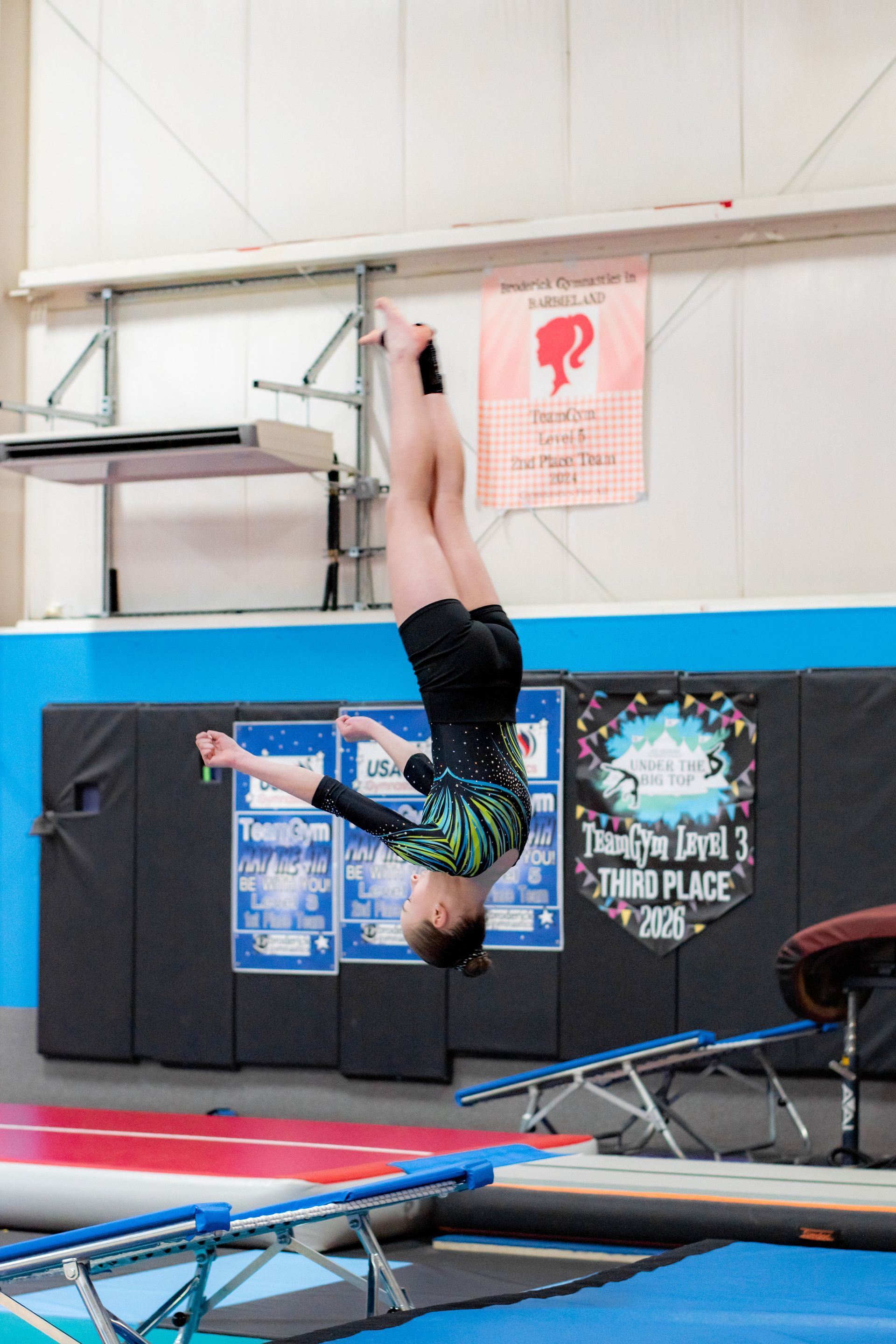 Gymnast hanging upside down on a trampoline indoors, wearing a green top and black shorts.
