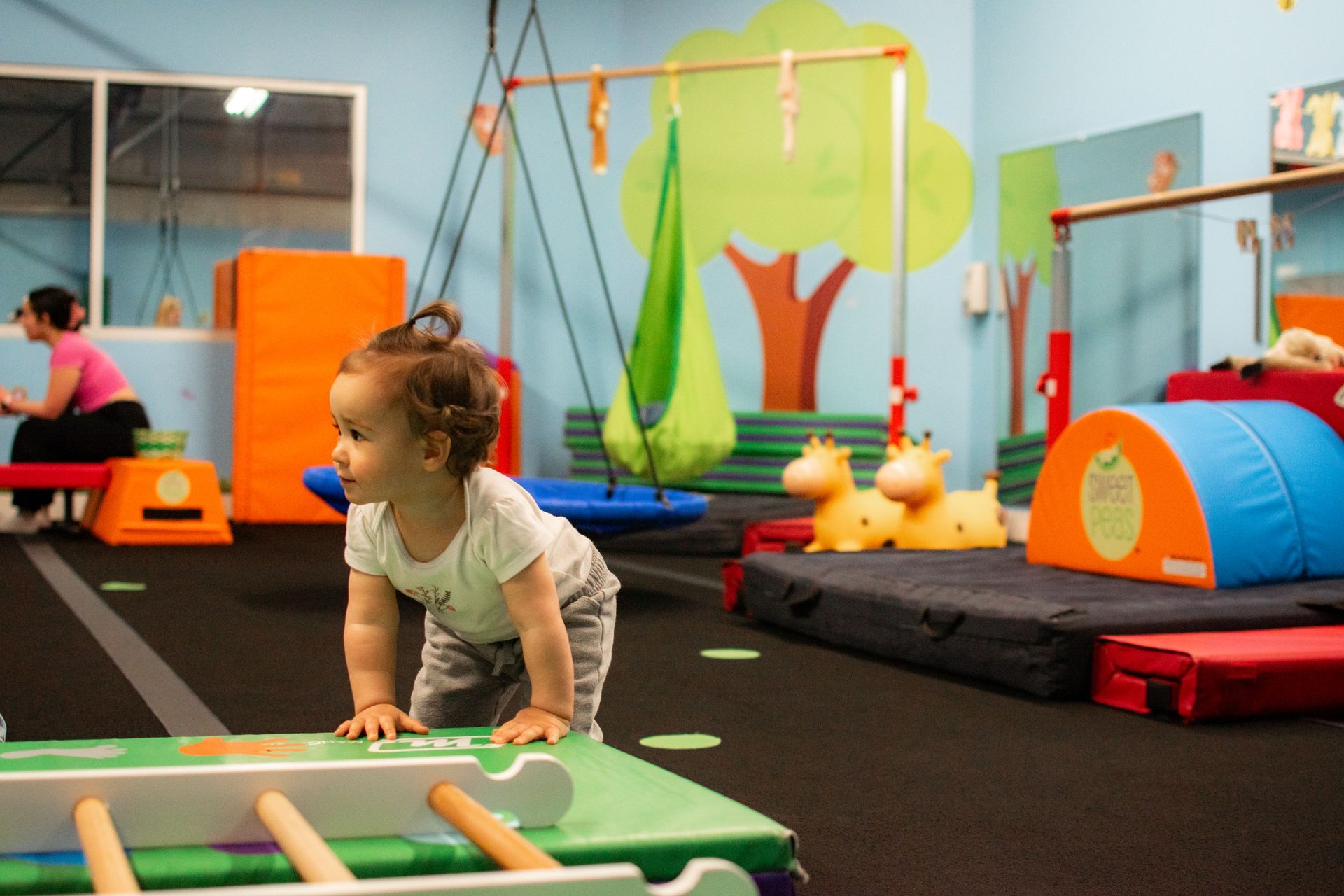 Toddler climbing on gym equipment in a colorful indoor playroom with mats, tunnels, and swings