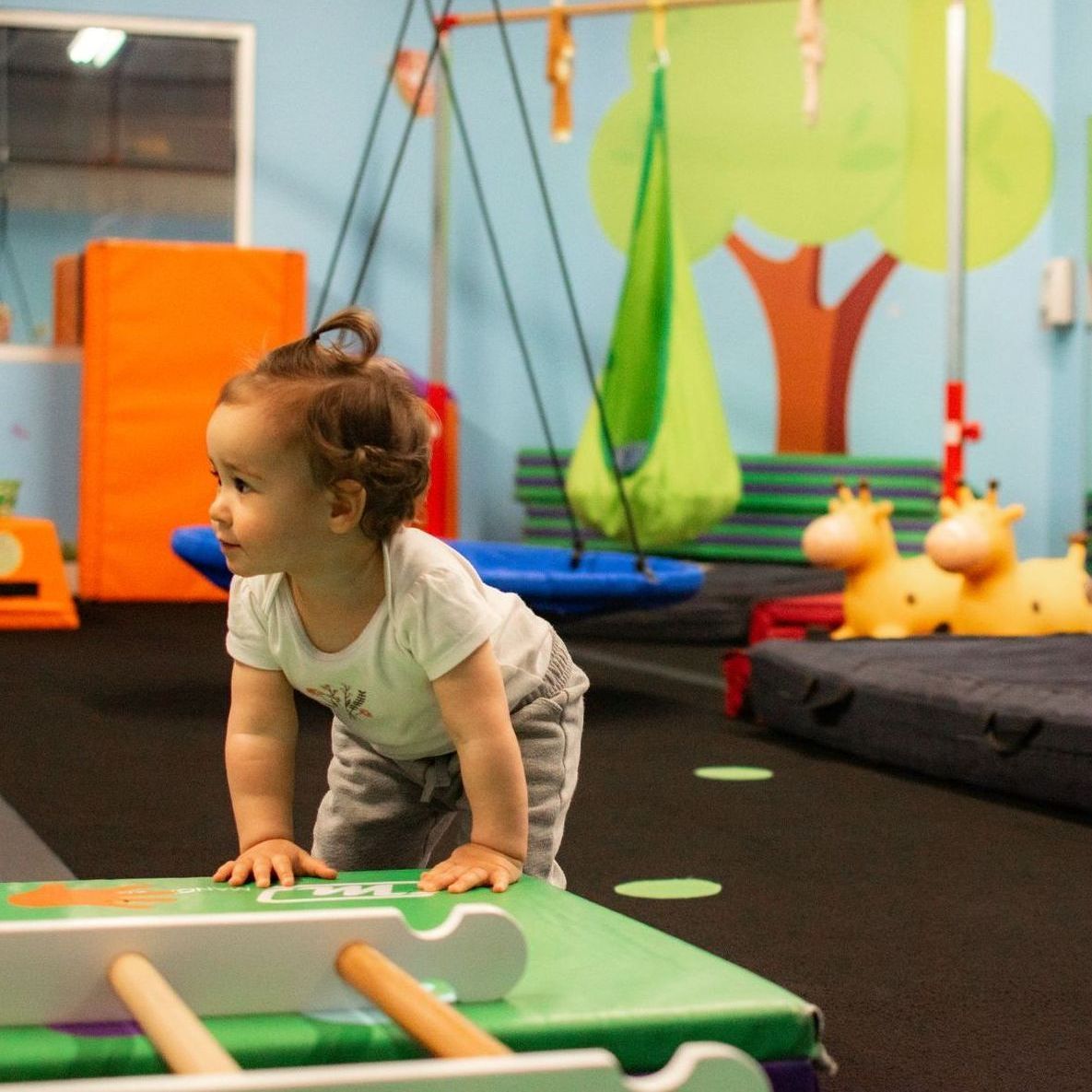 Toddler crawling on a green play platform in a colorful indoor playroom with toys and padded mats
