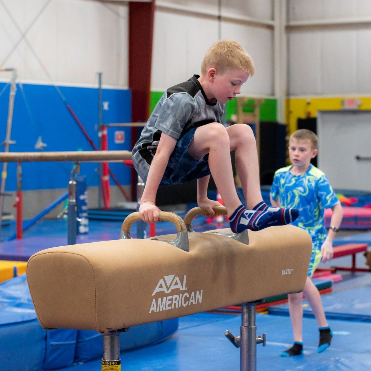 Child on pommel horse in a gym, with another child spotting nearby.