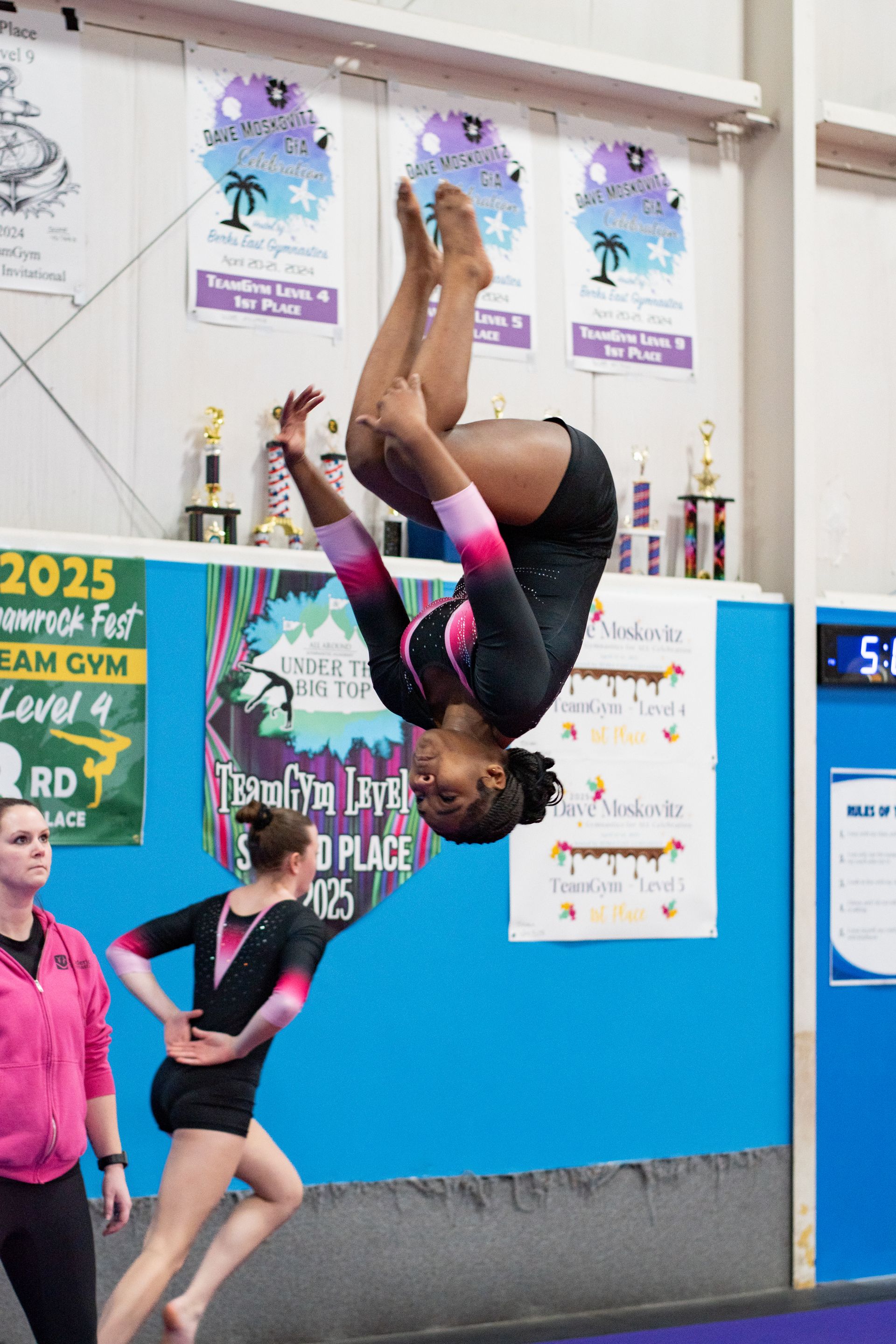 Gymnast mid-backflip indoors, upside down near the wall, with trophies and posters in the background