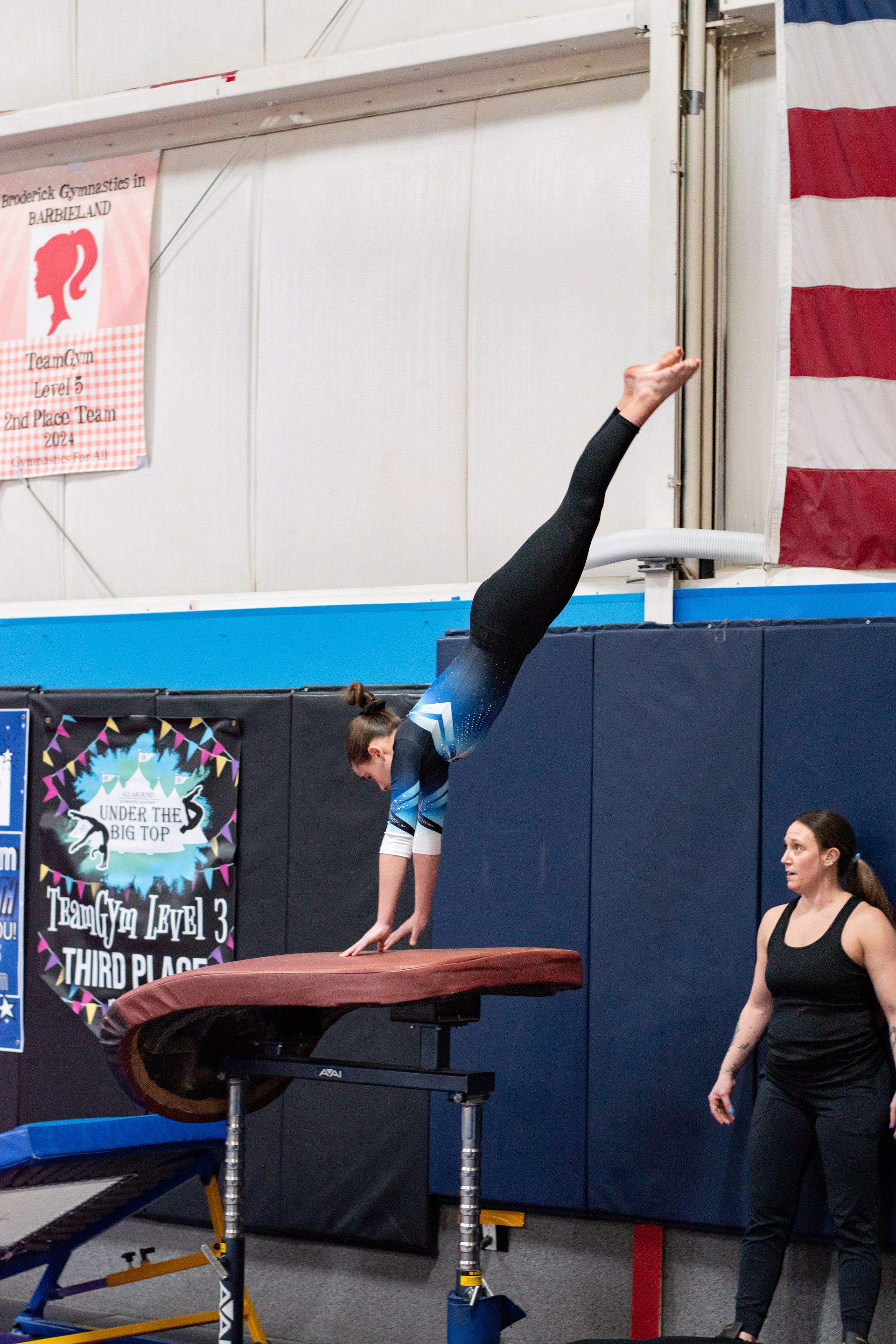 Gymnast balancing on one hand atop a vault table in an indoor gym, with a coach nearby
