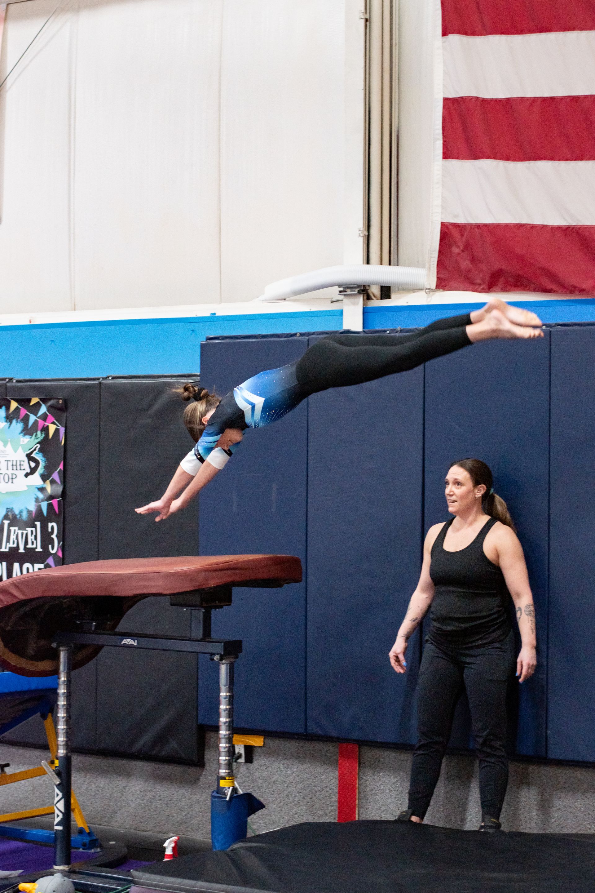 Gymnast mid-air over a vault while a coach stands nearby in a gymnasium