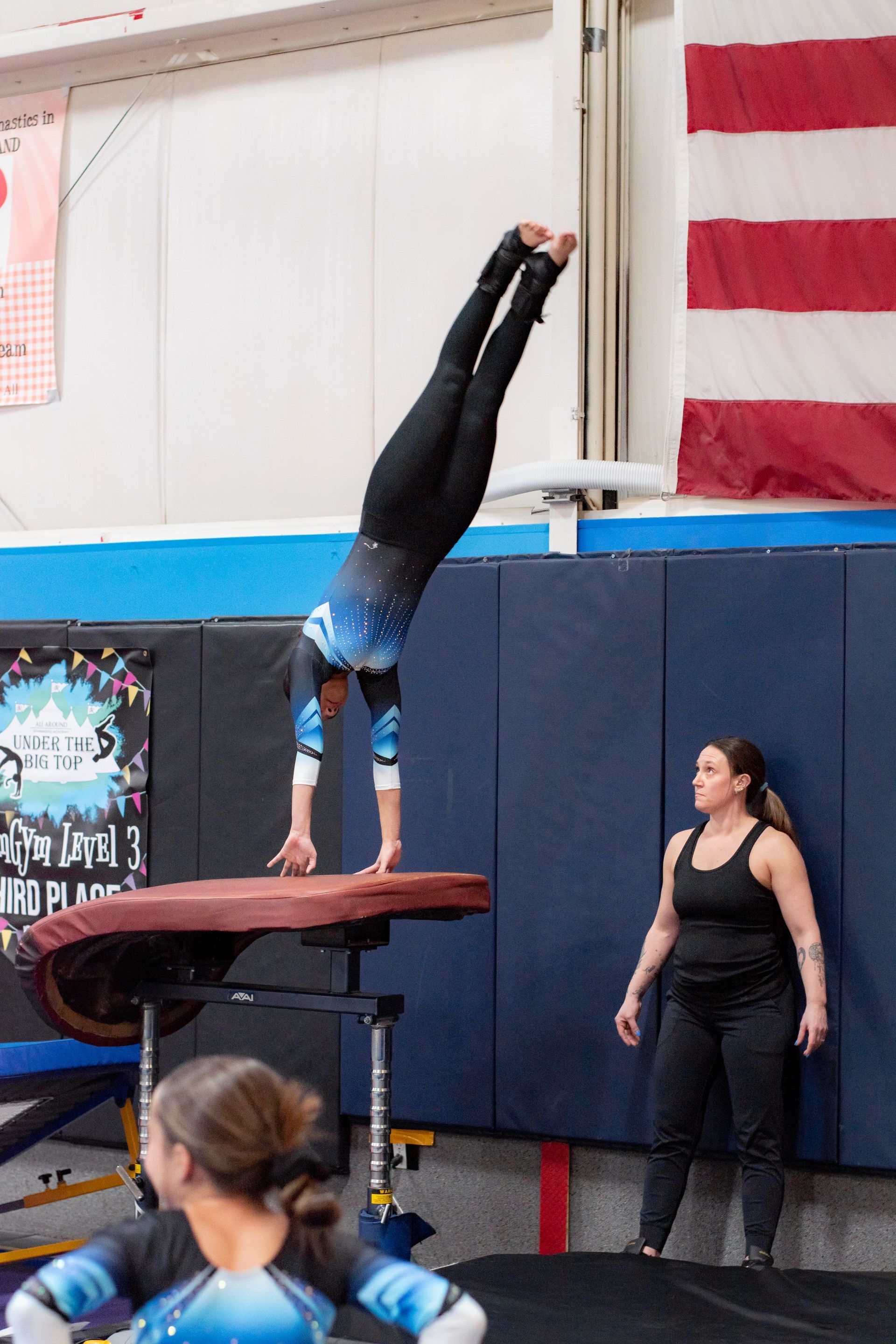 Gymnast doing a handstand on a balance beam while a coach watches nearby in a gym.