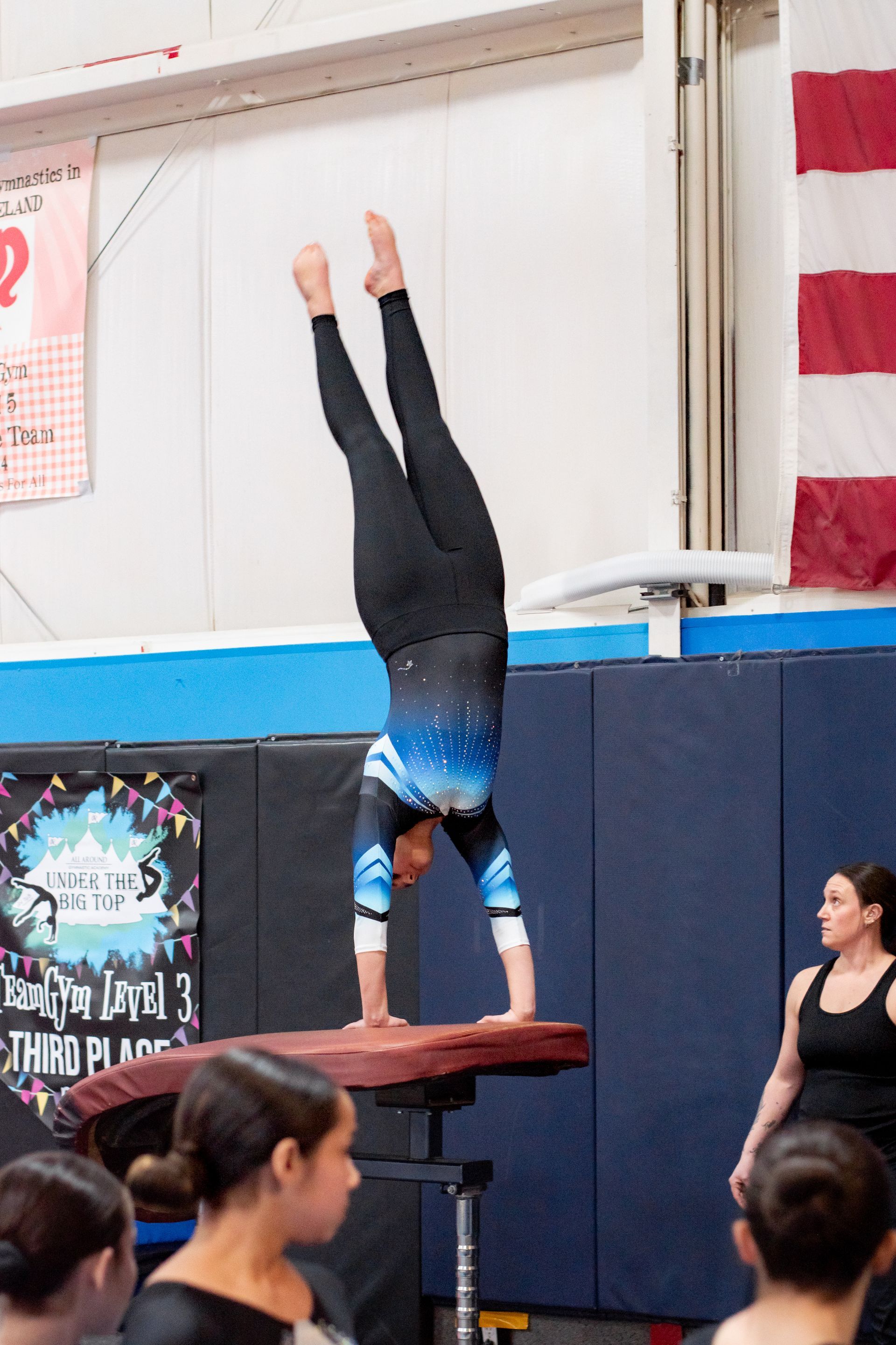 Gymnast holding a handstand on a vault table during an indoor performance, with spectators nearby