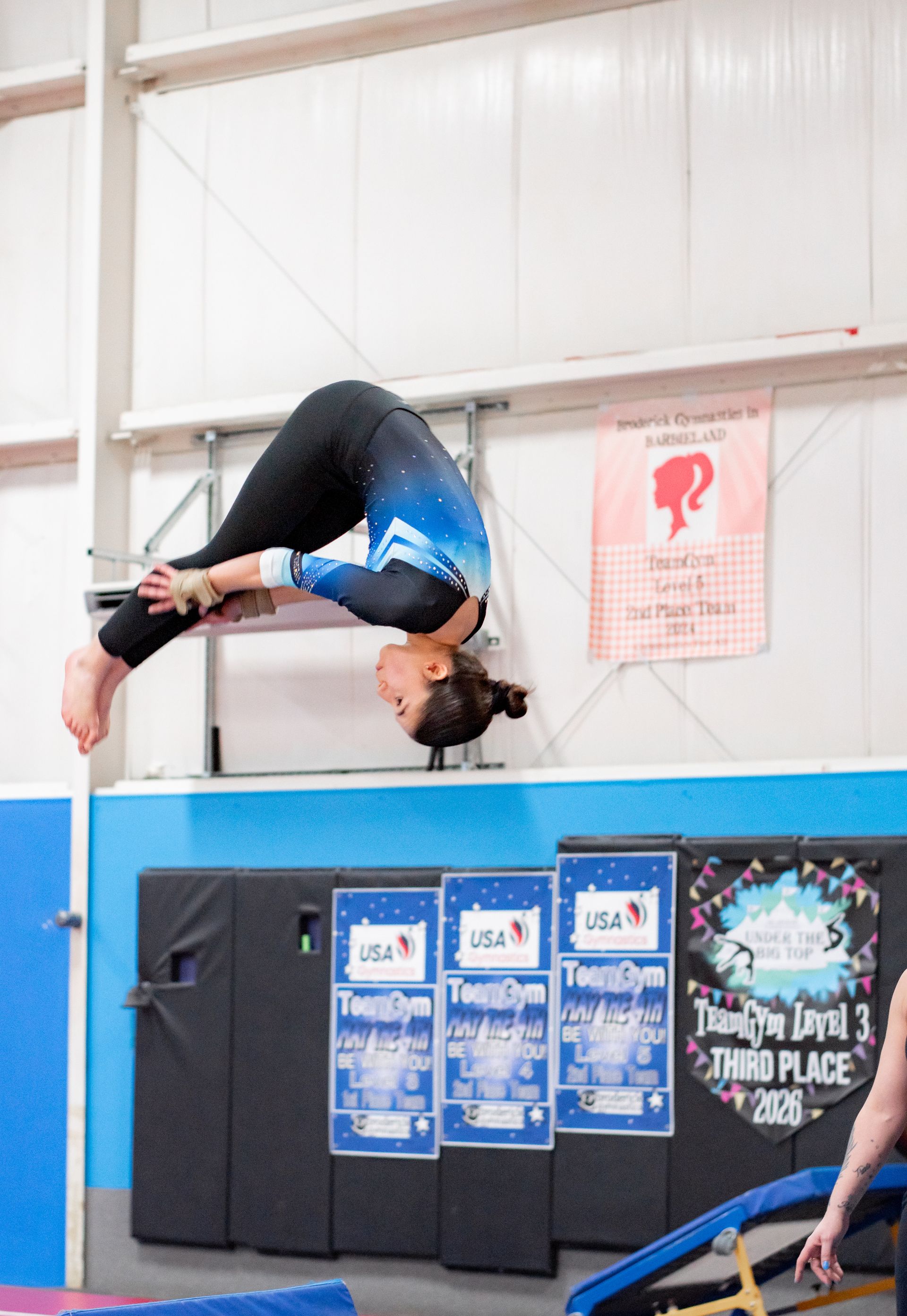 Gymnast mid-backflip over a foam pit in an indoor gym, with blue mats and wall banners nearby