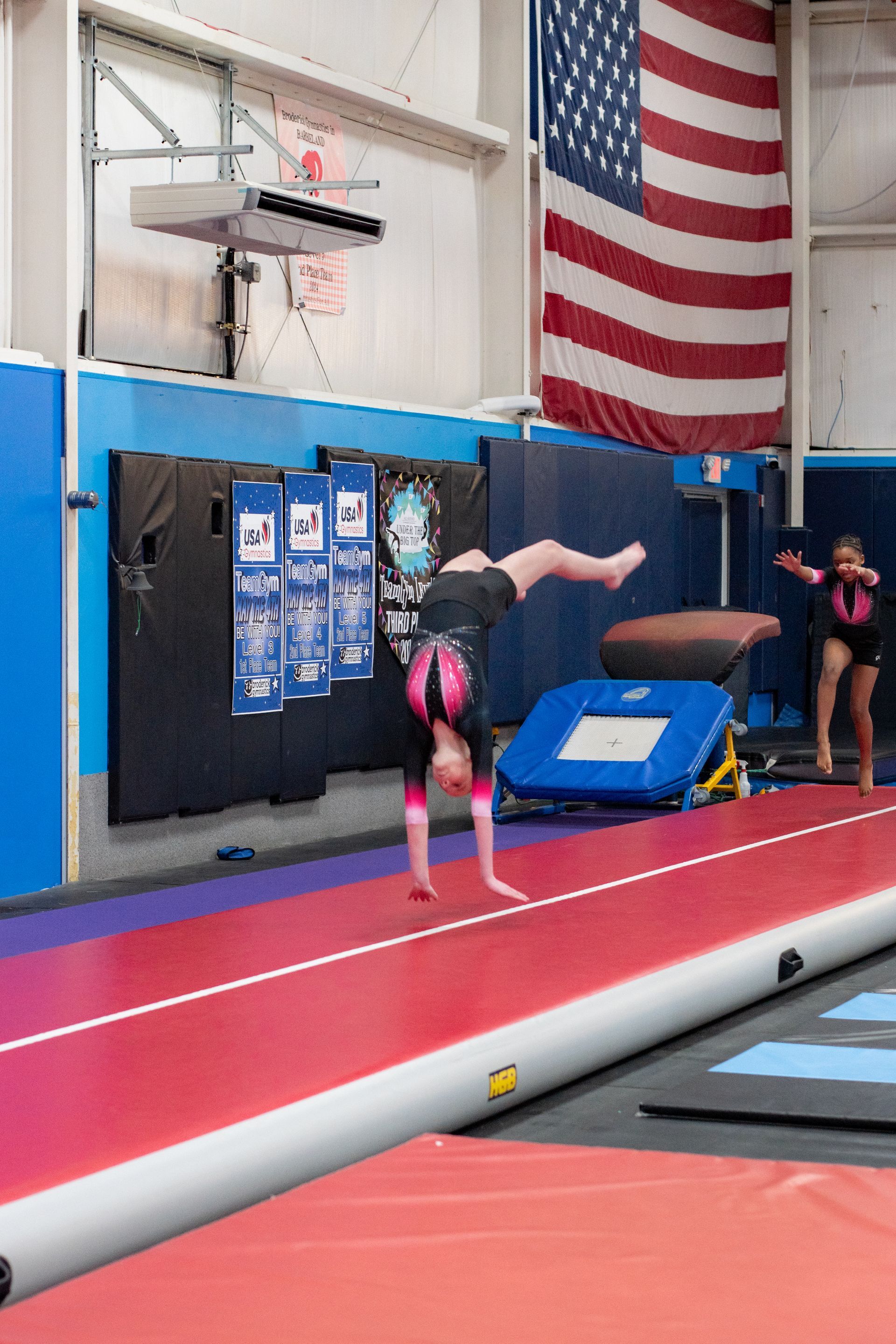 Gymnast mid-flip on a red runway in an indoor gym, with blue mats and an American flag in the background