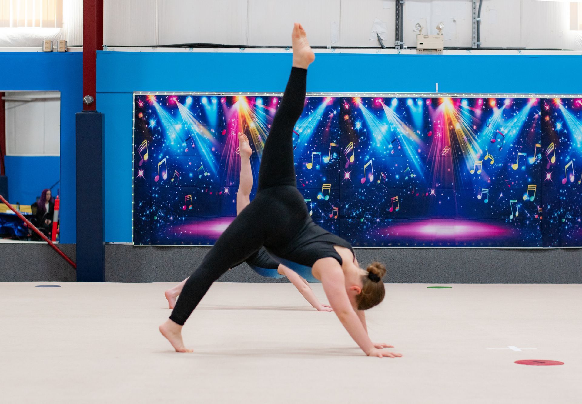 Gymnast in black holds a one-arm balance on a mat in an indoor gym.