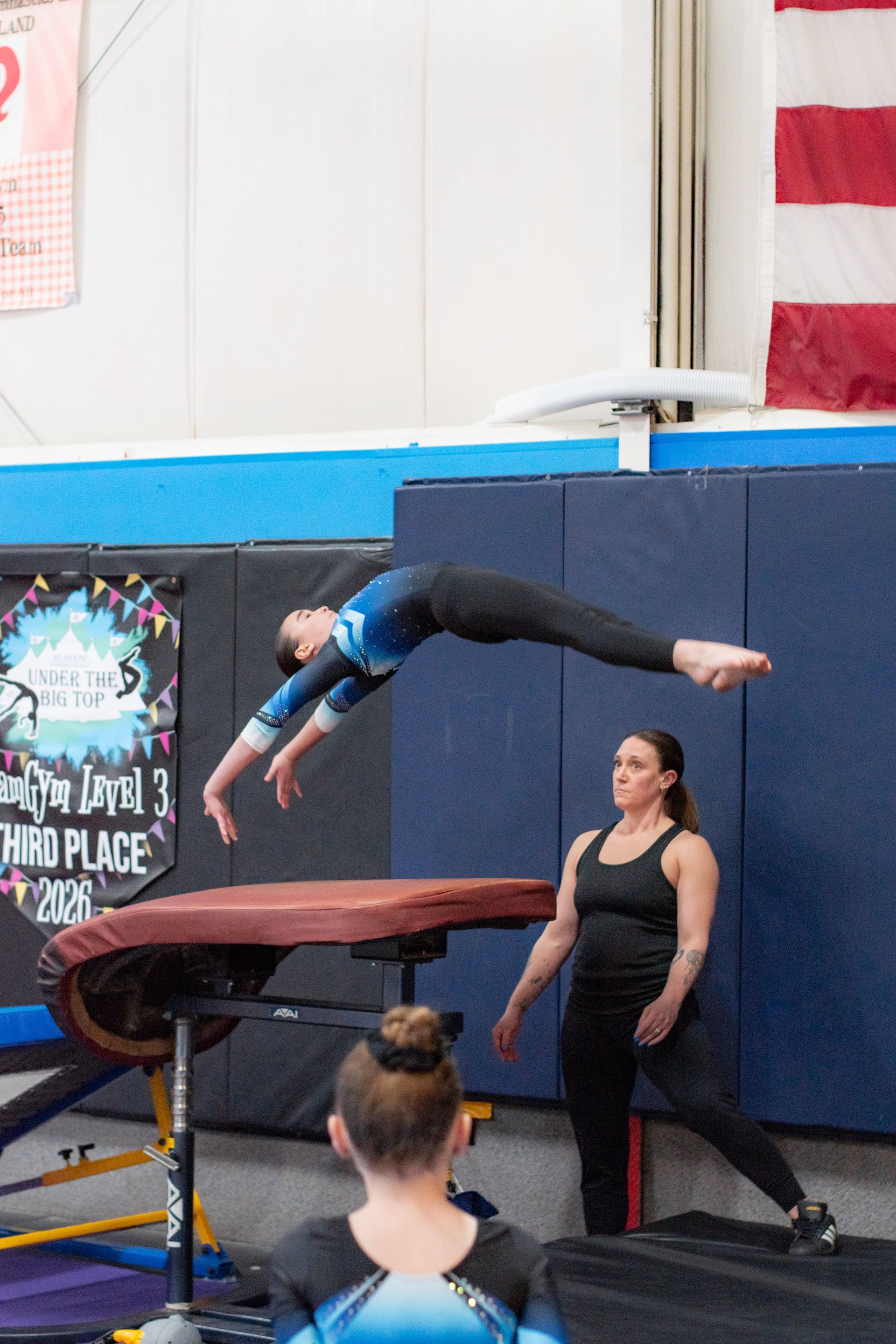 Gymnast performing a back handspring over a vault, with a coach spotting in an indoor gym.
