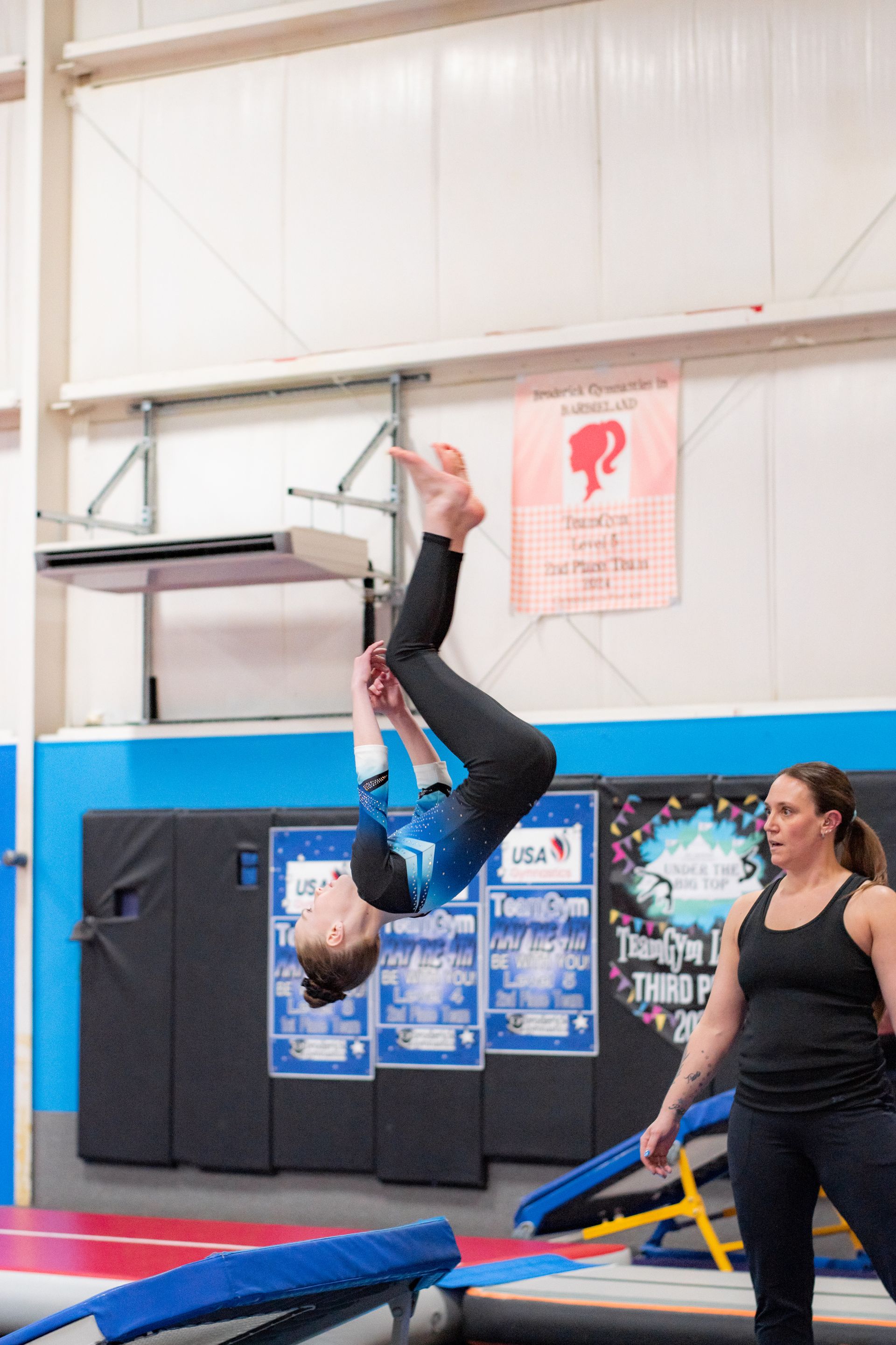 Gymnast upside down on trapeze while another woman stands nearby in a gym