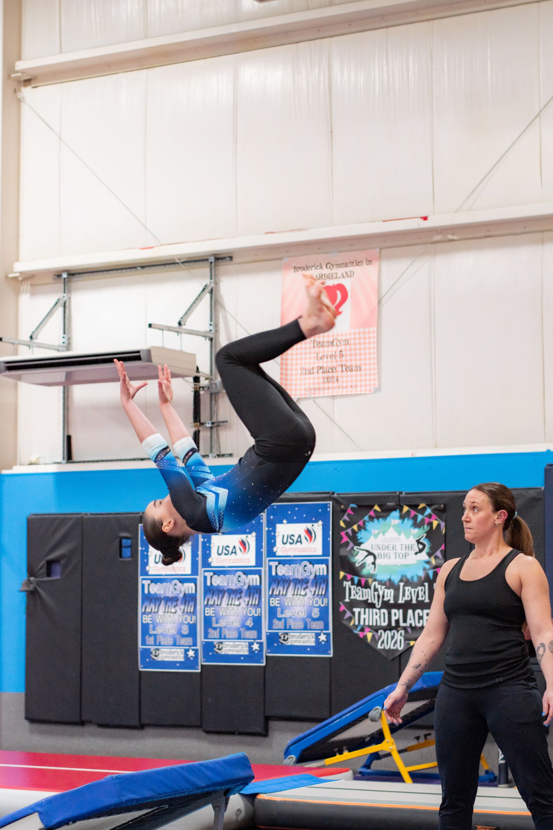 Gymnast flipping backward on a trampoline while a coach watches nearby