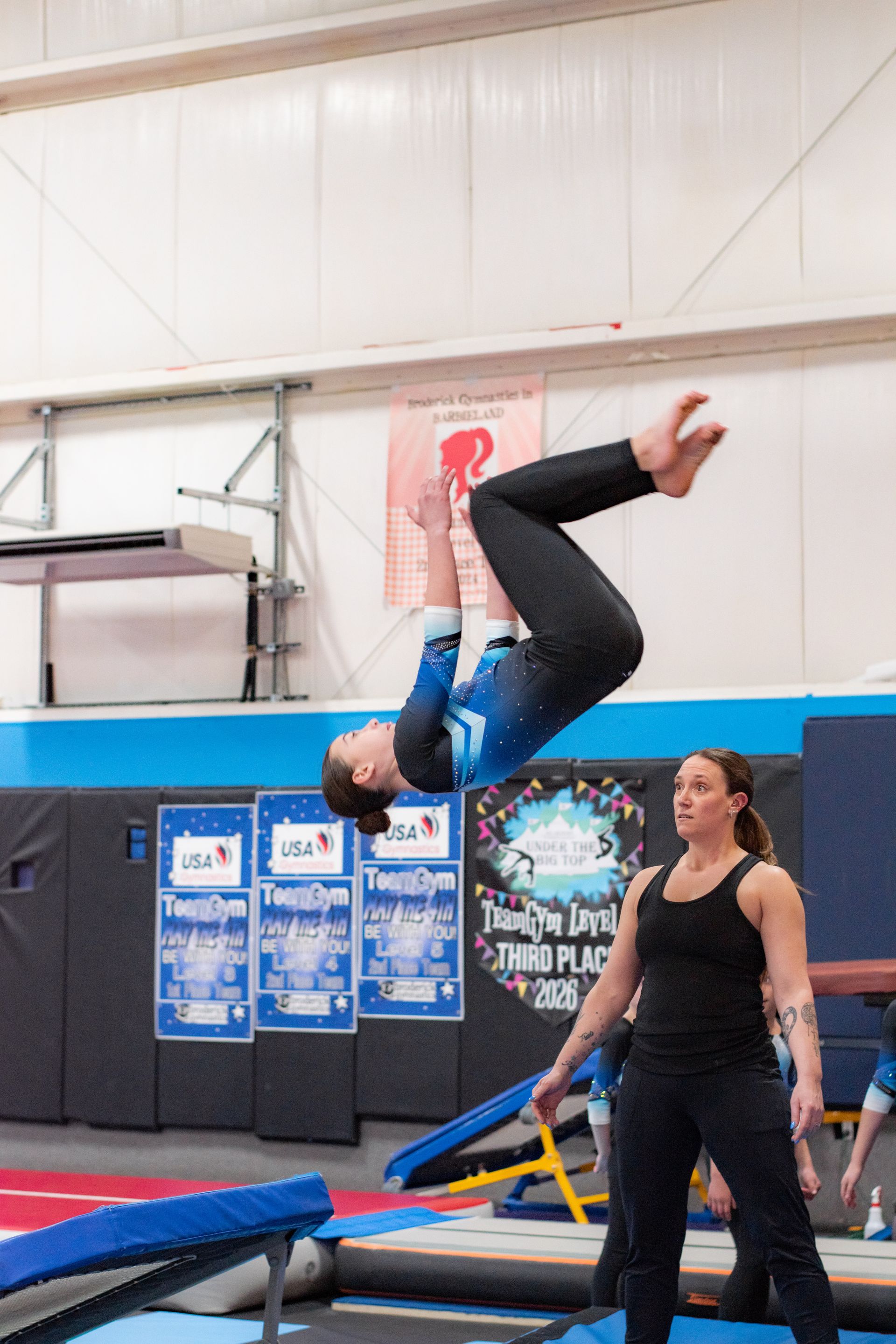Gymnast flipping backward on a trampoline in an indoor gym while a coach watches nearby