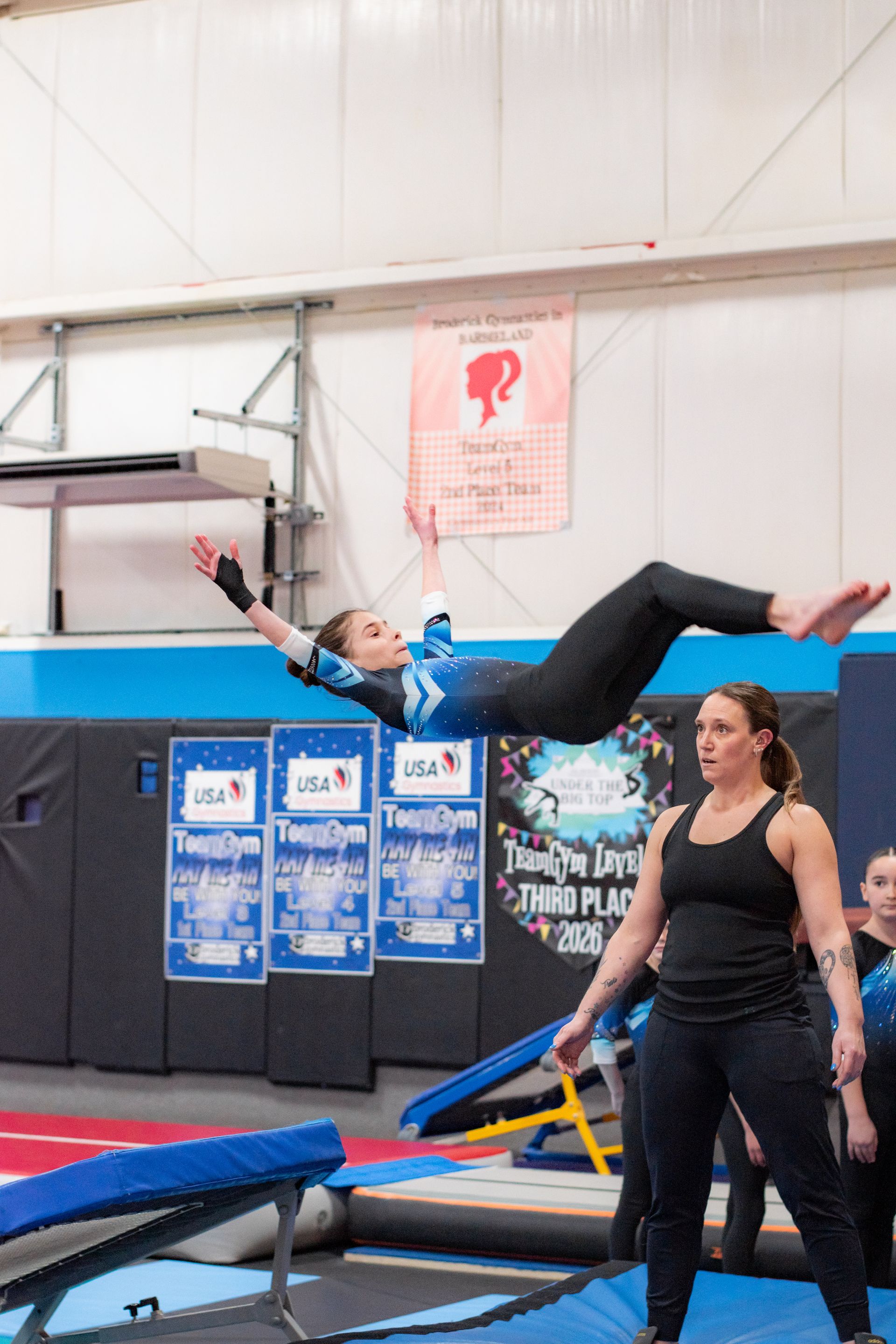 Gymnast midair above a trampoline while a coach watches nearby in an indoor gym