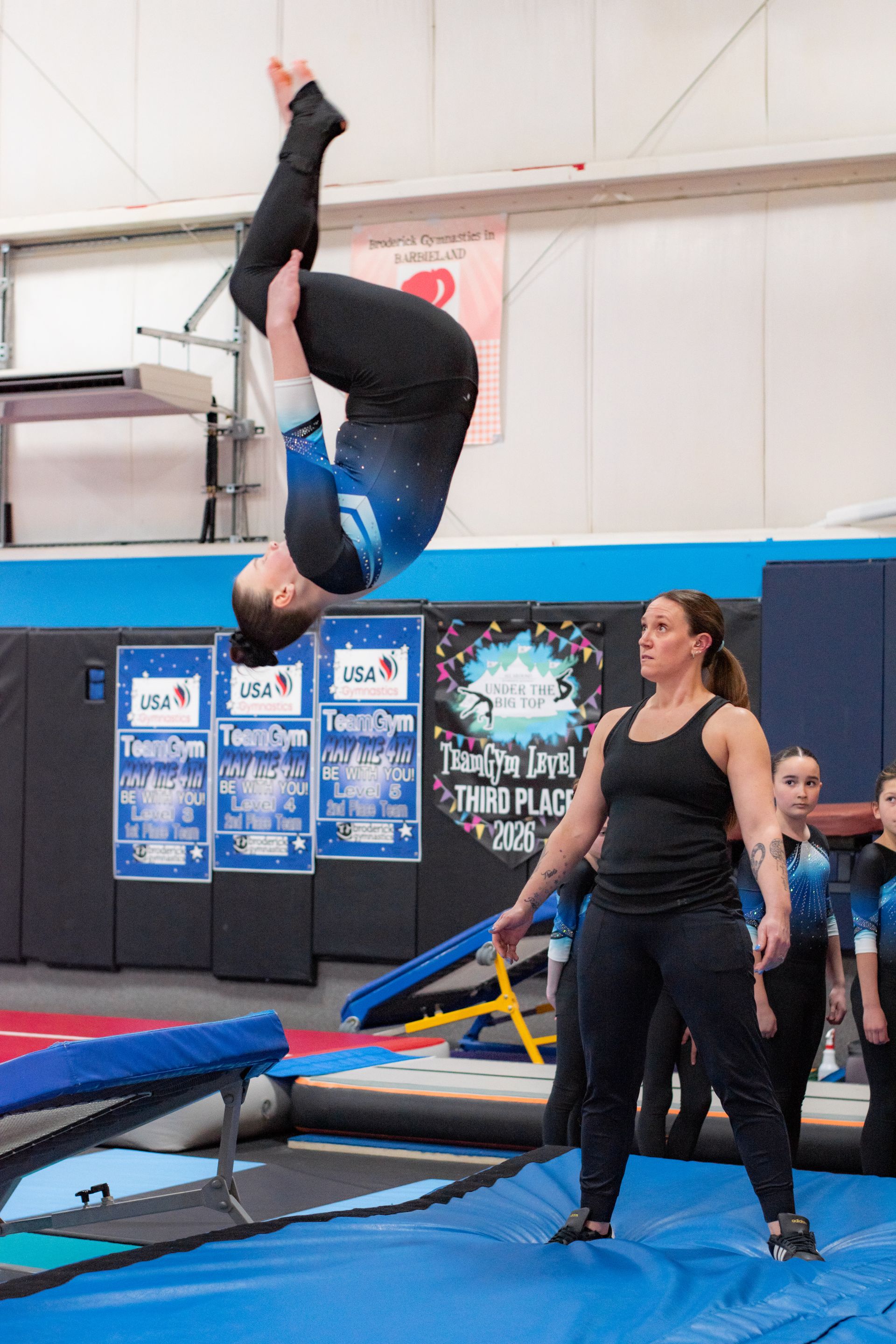 Gymnast mid-backflip on a trampoline while a coach watches nearby in an indoor gym