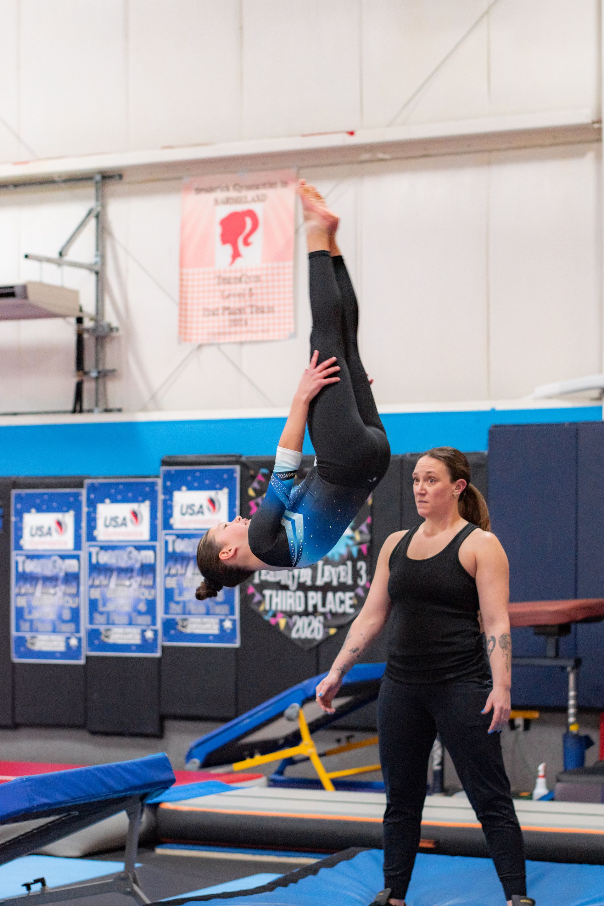 Gymnast upside down in a handstand on a balance beam, with a coach standing nearby in a gym.