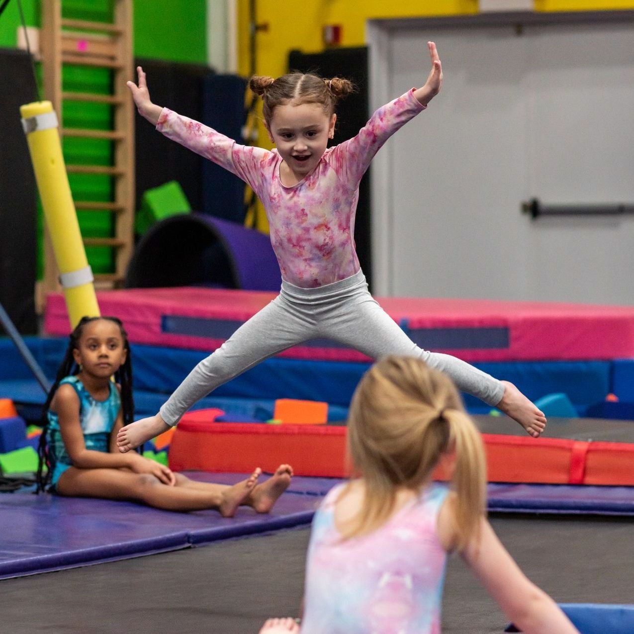 Child doing a split jump in a gymnastics gym, with other children and colorful mats nearby.