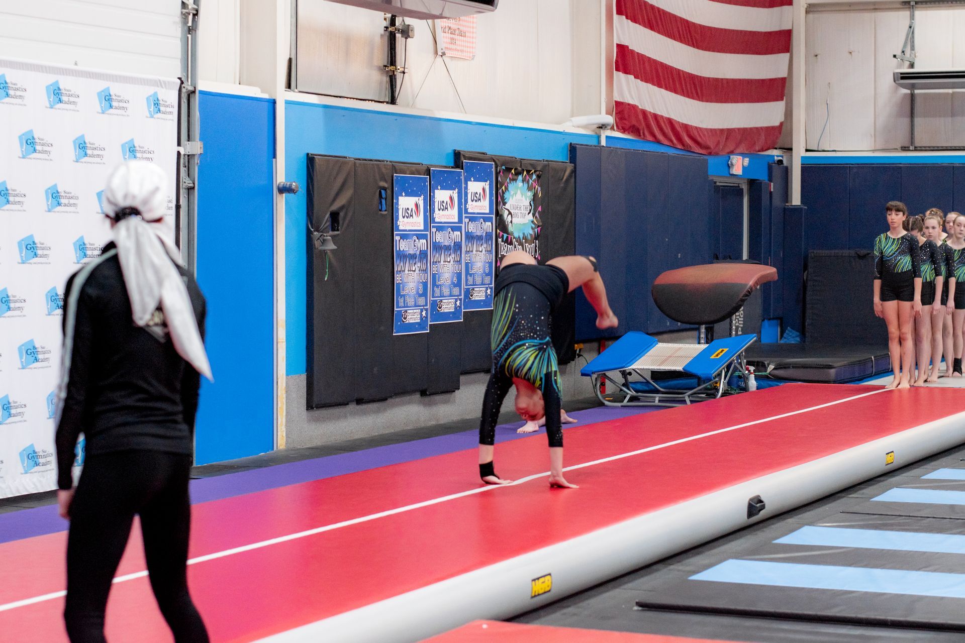 Gymnast performing a vault in a gym, with teammates watching nearby and an American flag in the background