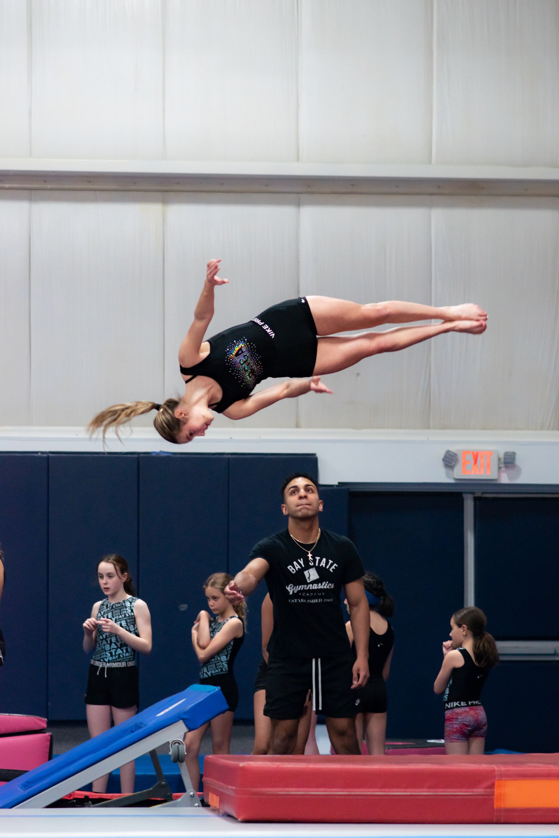 Gymnast midair in a backflip above a blue springboard, with spectators watching in a gym.