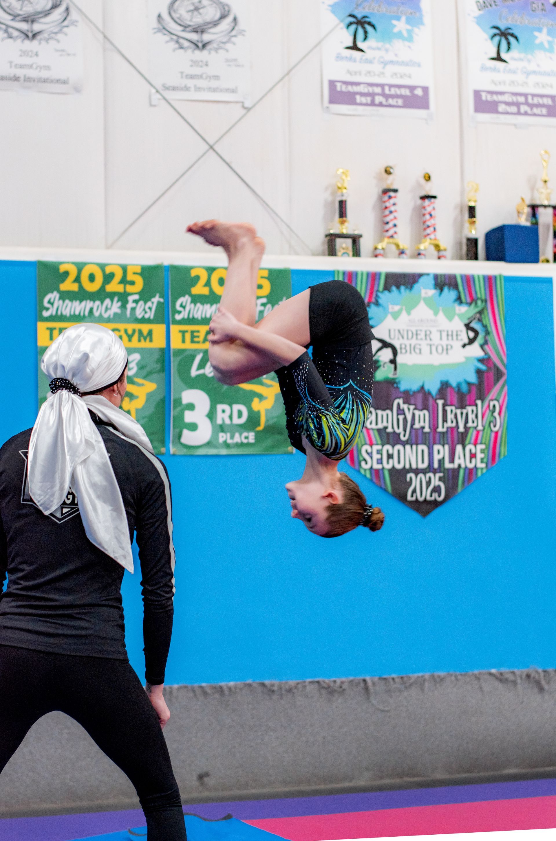 Gymnast mid-flip in front of a blue competition wall with award posters and trophies