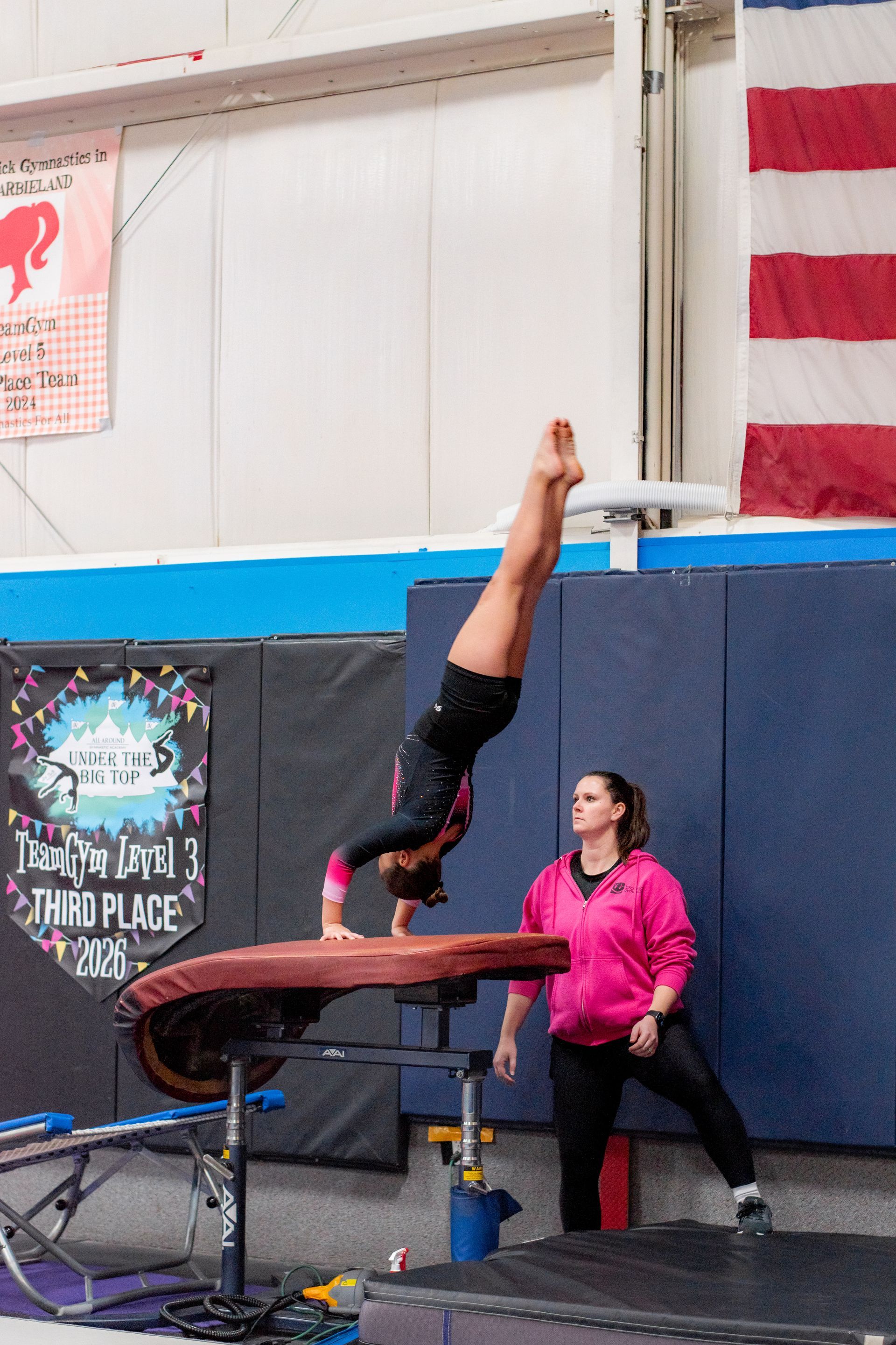 Gymnast performing a handstand on a vault, with a coach spotting in an indoor gym.