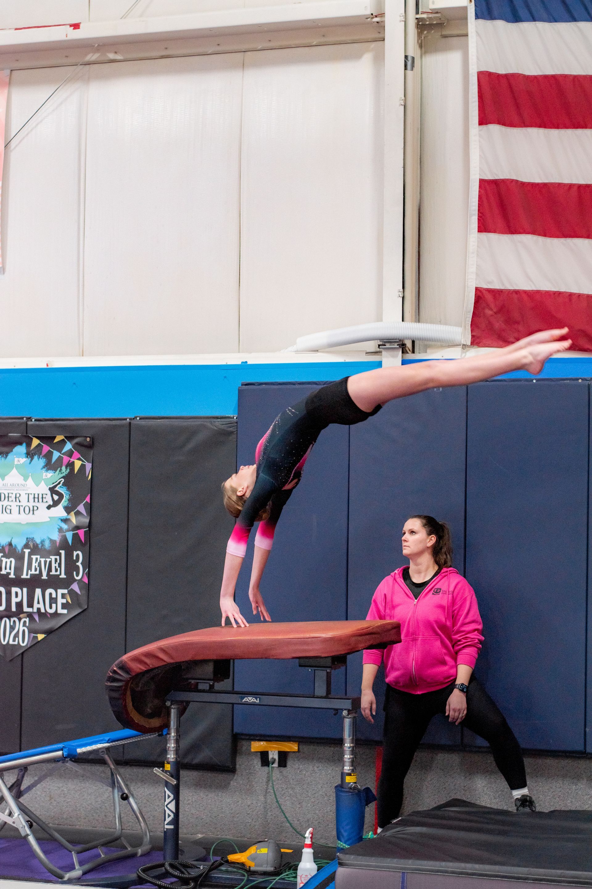 Gymnast balancing in a handstand on a padded vault table in a gym, with a coach spotting nearby.