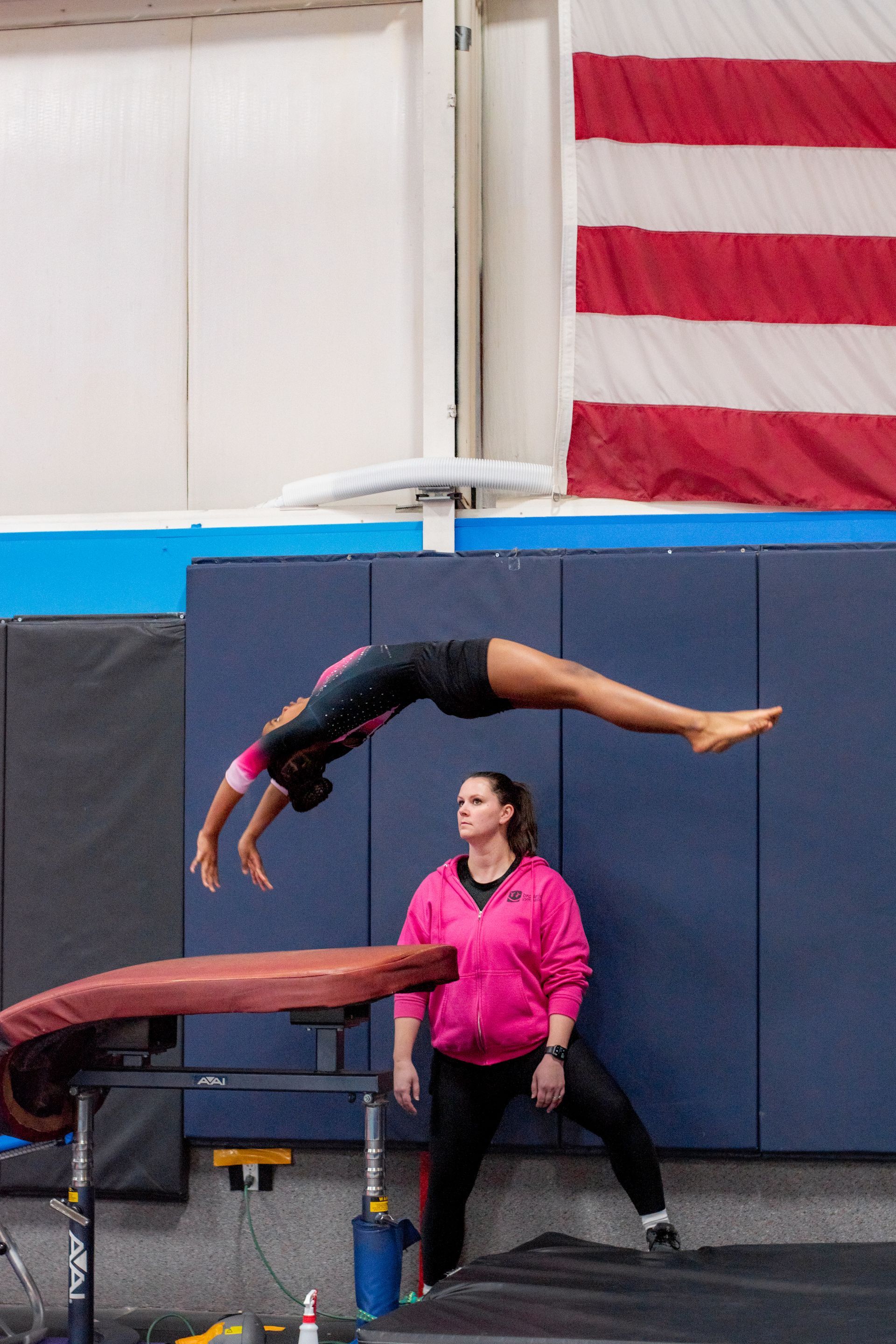 Gymnast mid-backflip over a vault while a coach spots her, with a U.S. flag in the background