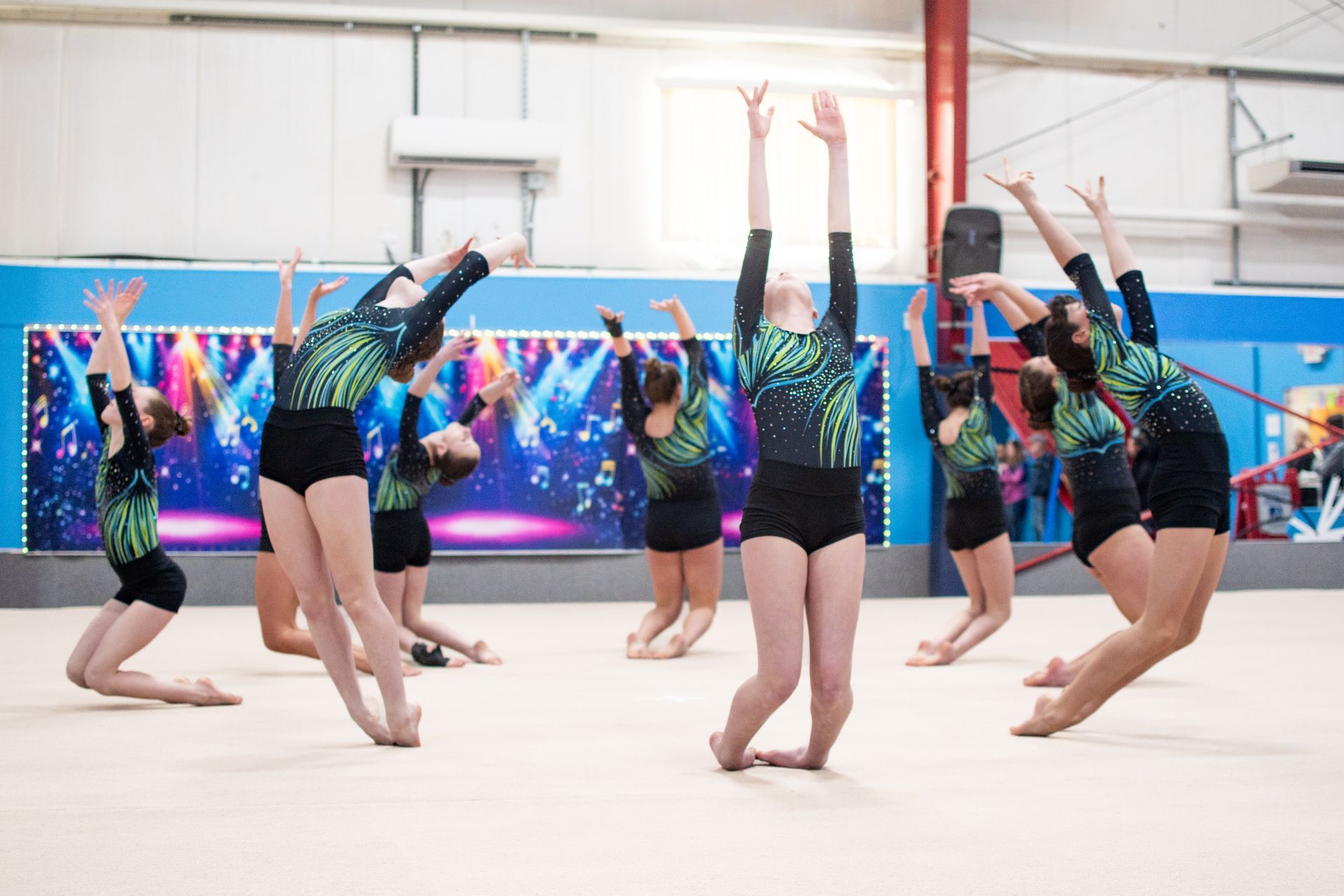 Gymnasts performing synchronized back handsprings in a gymnasium, with a coach and digital screens in the background