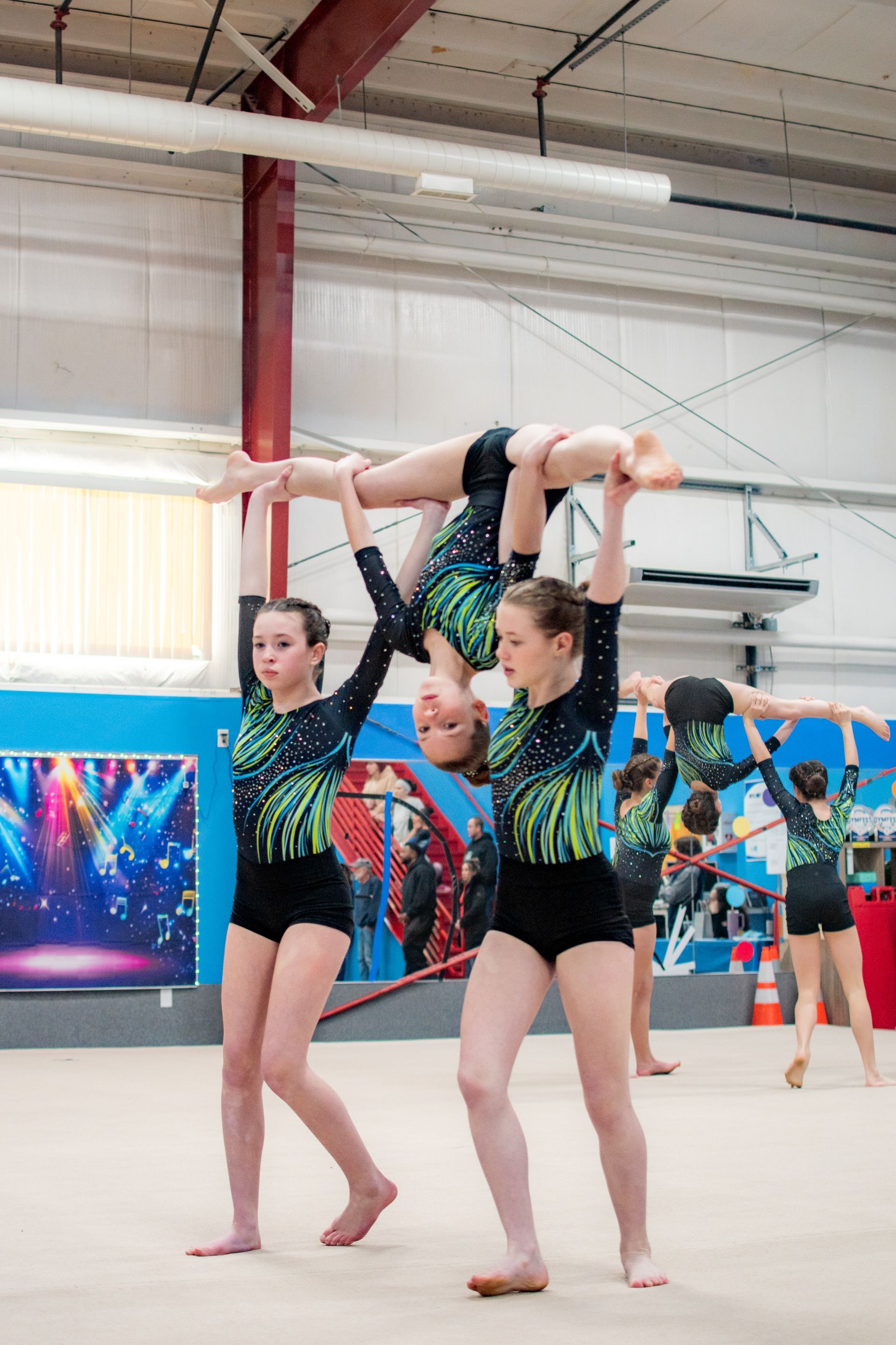 Three gymnasts in black and green leotards perform an acrobatic pose in a gymnasium