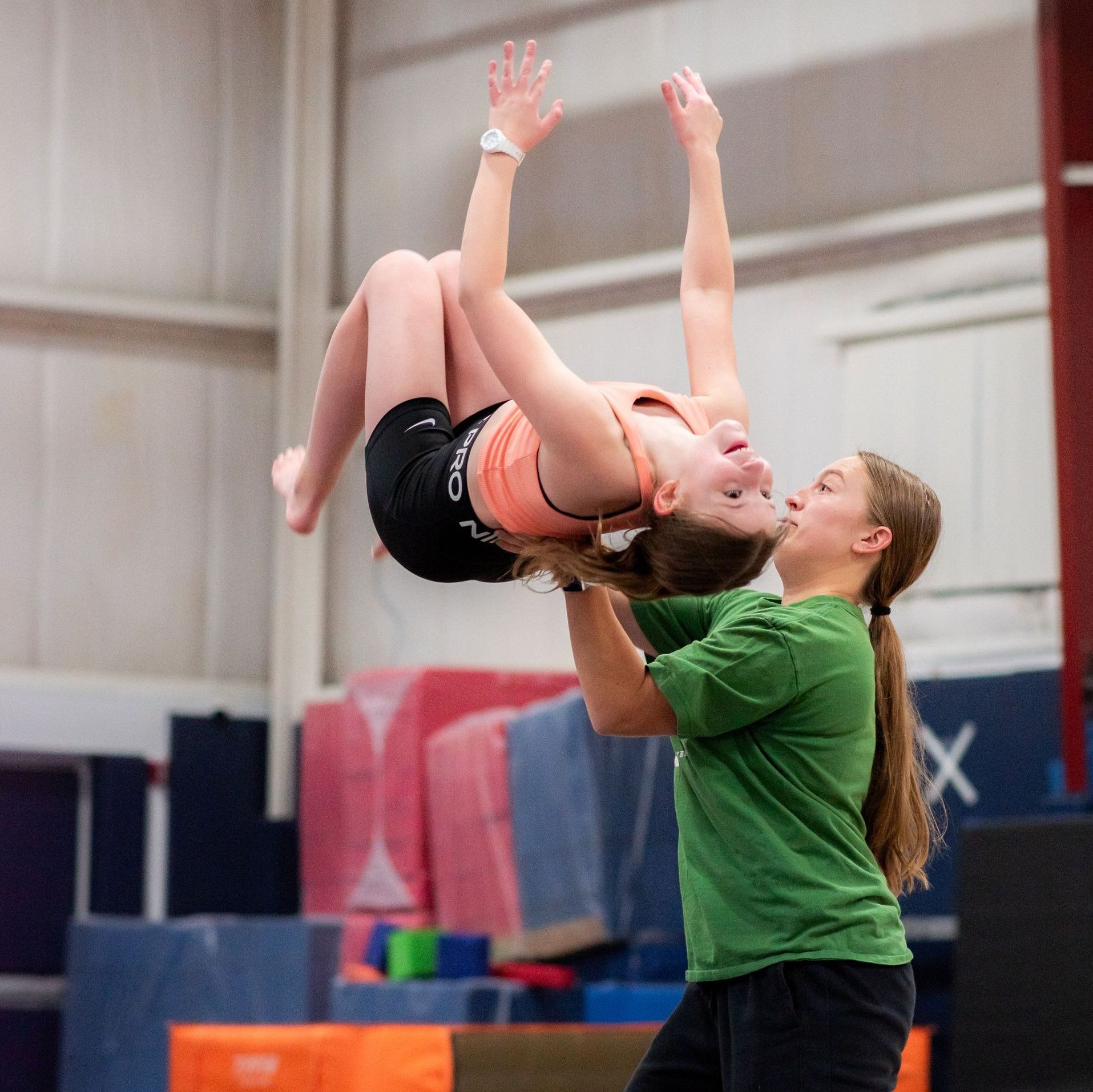 Gymnast being lifted in a gym, arms raised, by a coach in a green shirt.