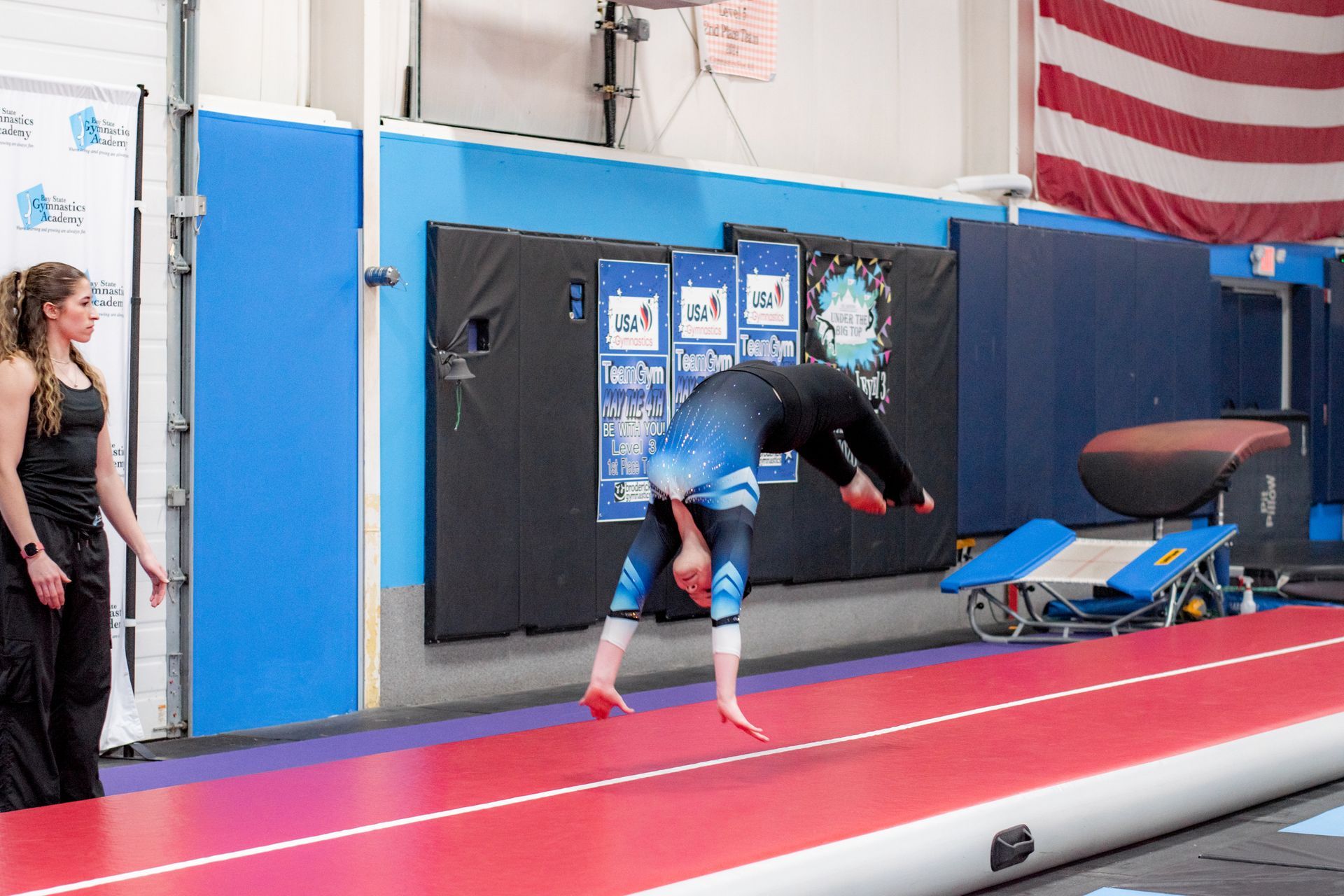 Gymnast mid-backflip on a red runway in a gym, with blue mats and a spotter nearby.