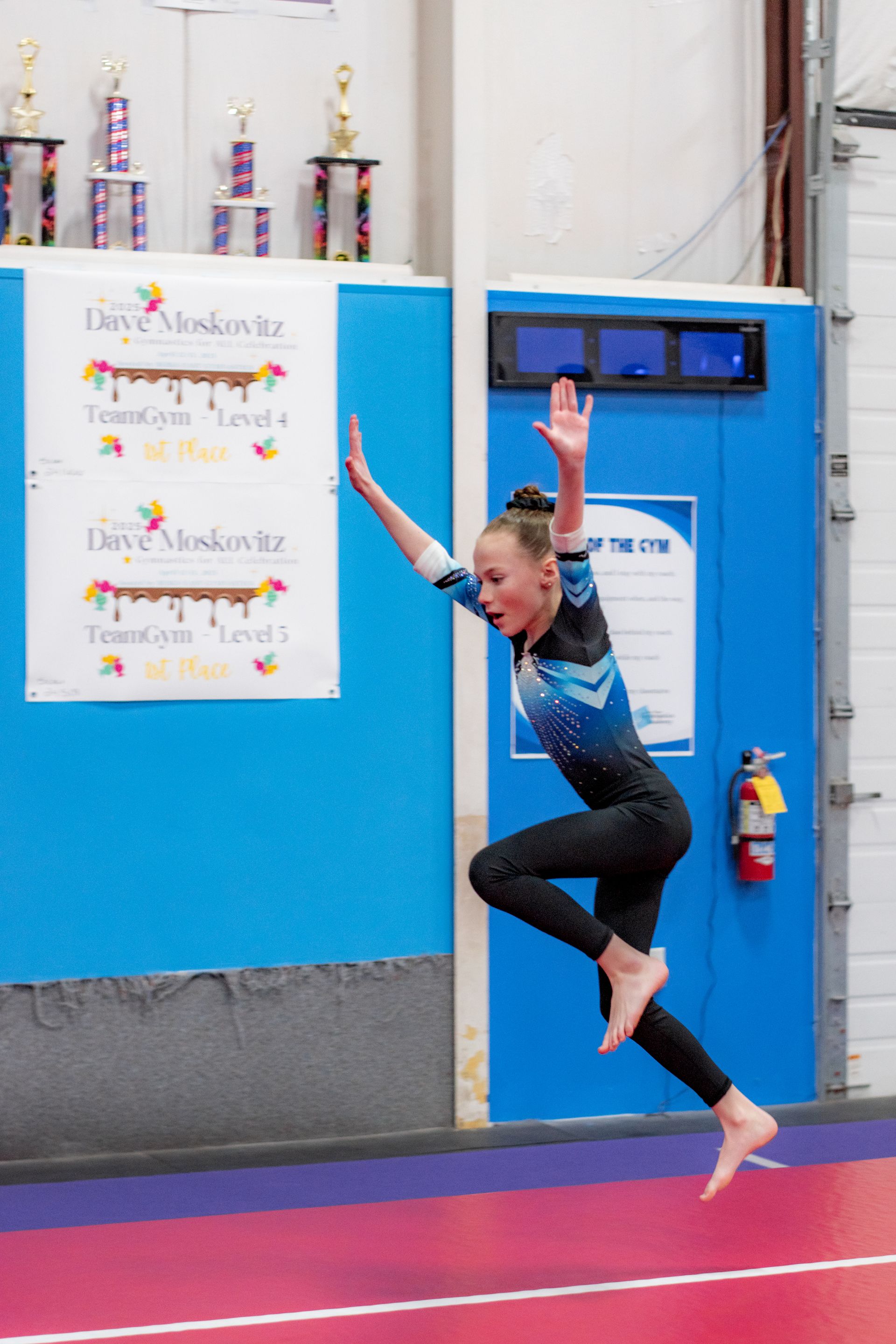 Gymnast mid-leap in blue leotard at an indoor gymnastics gym, arms raised near a blue wall.