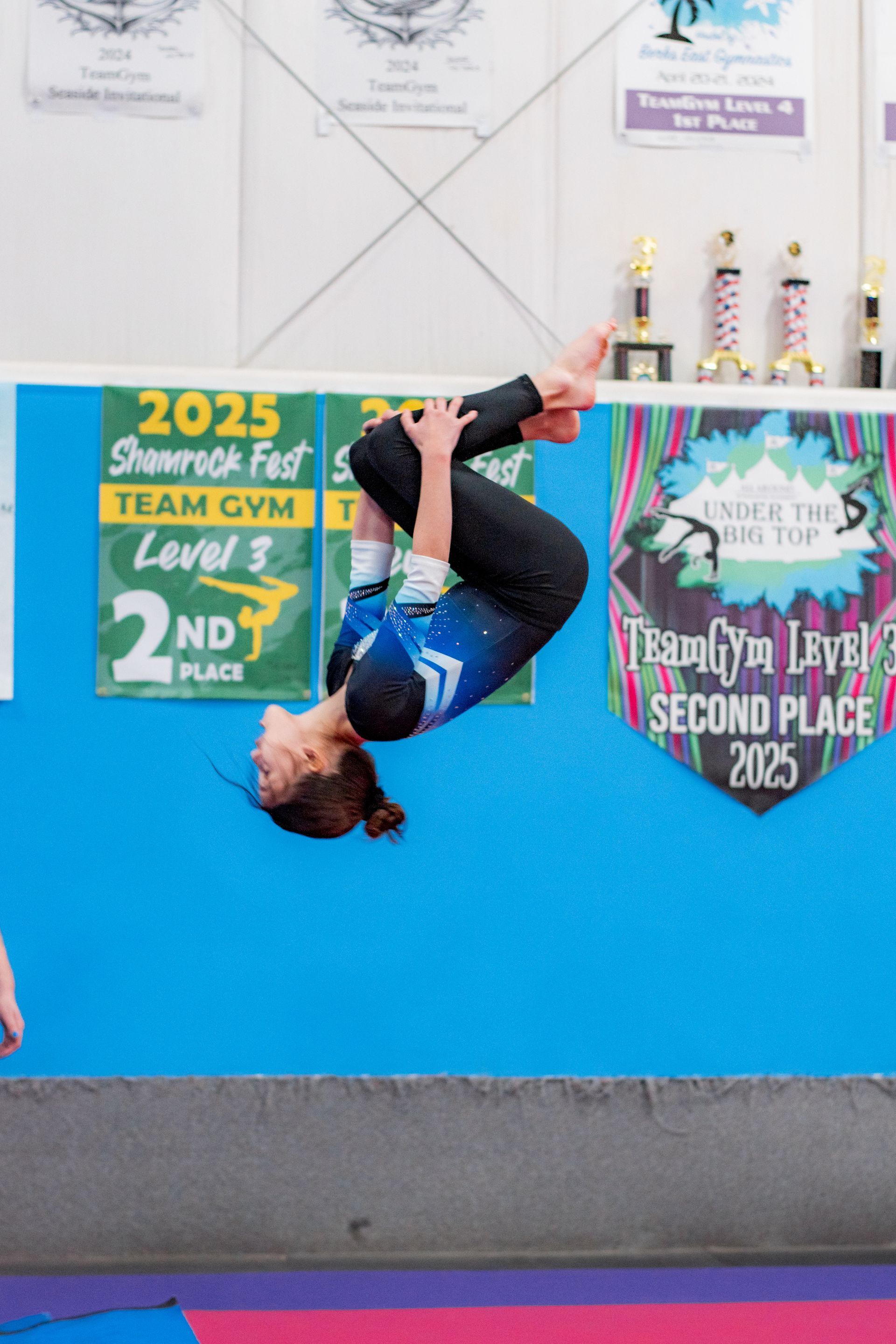 Dancer in blue shirt performing a backflip in a gym, with competition banners in the background.