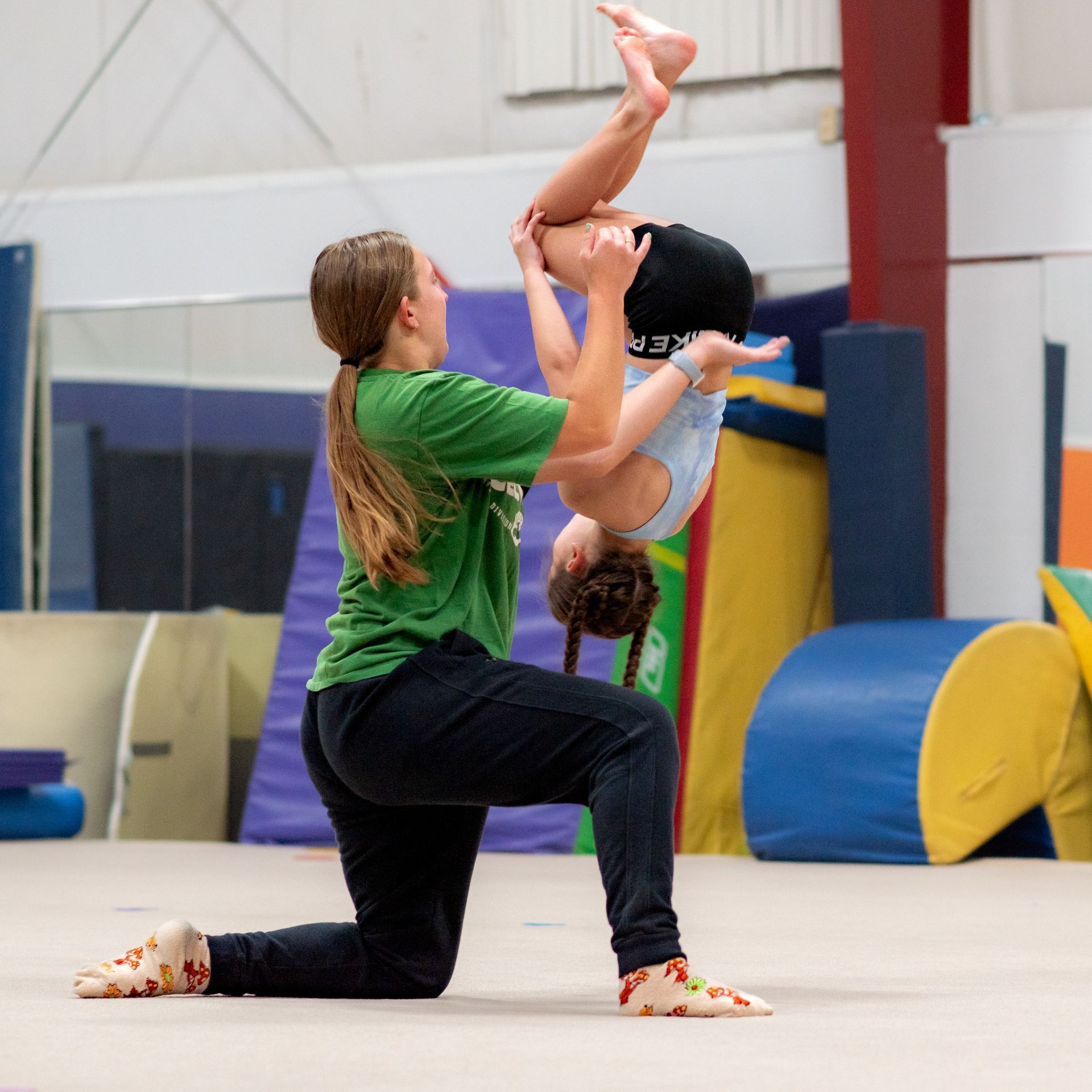 Gymnastics coach spotting a child mid-backflip in a gym, with colorful mats in the background