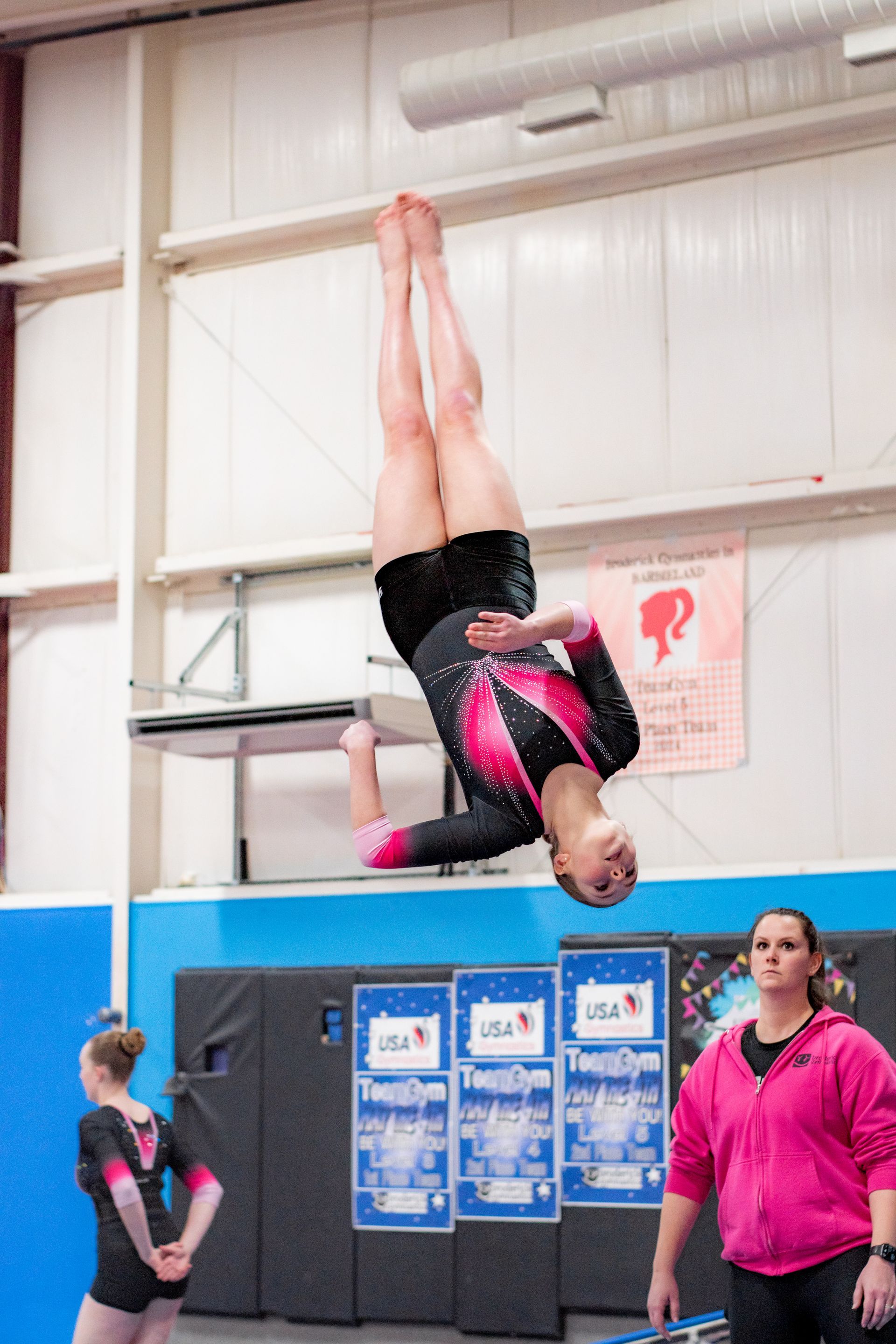 Gymnast upside down on high bar during training indoors, with coaches watching nearby.