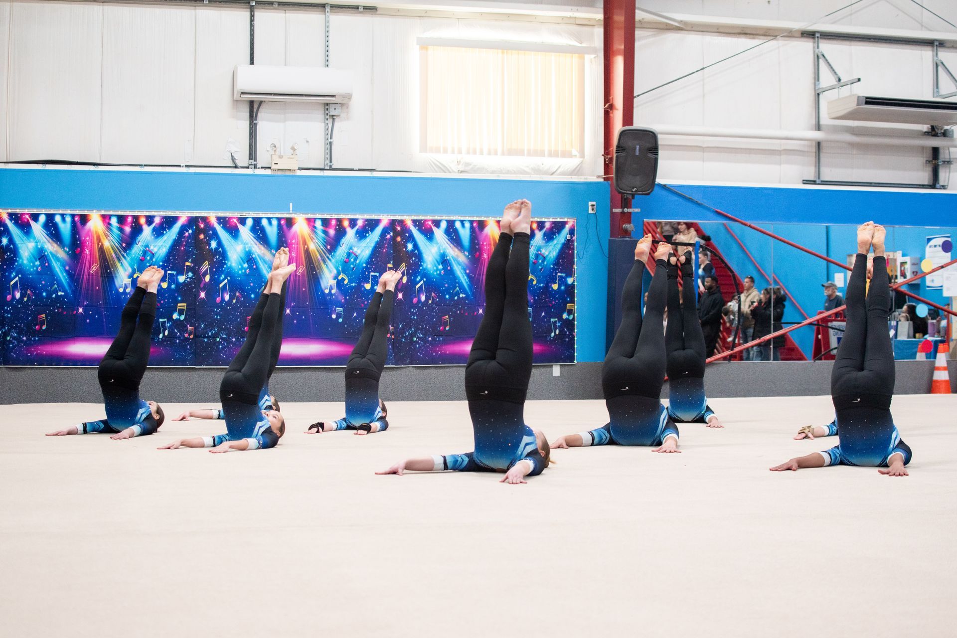 Gymnasts in blue practice synchronized headstands on a mat in an indoor gym.
