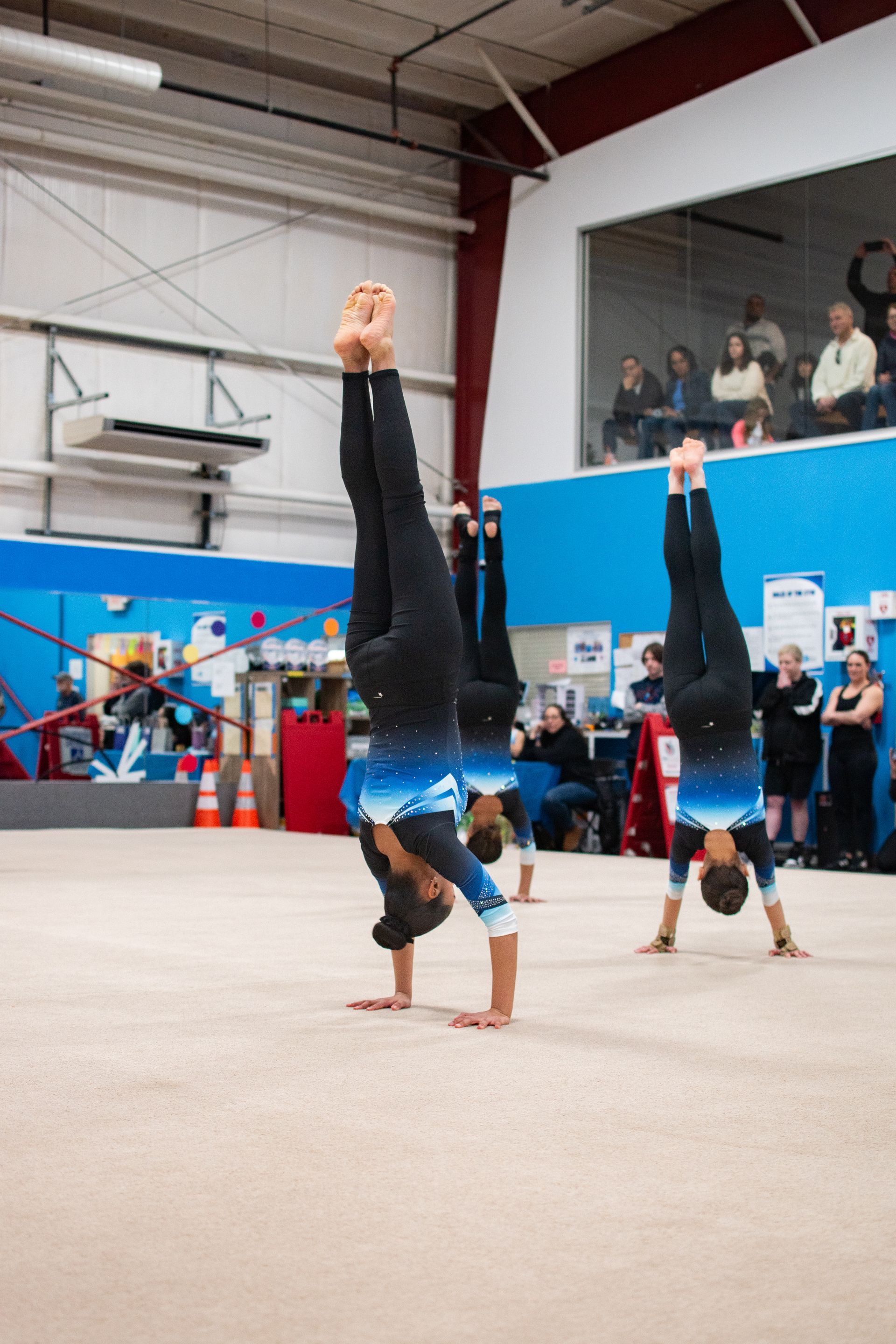 Two gymnasts in blue uniforms doing handstands on a gymnastics floor indoors