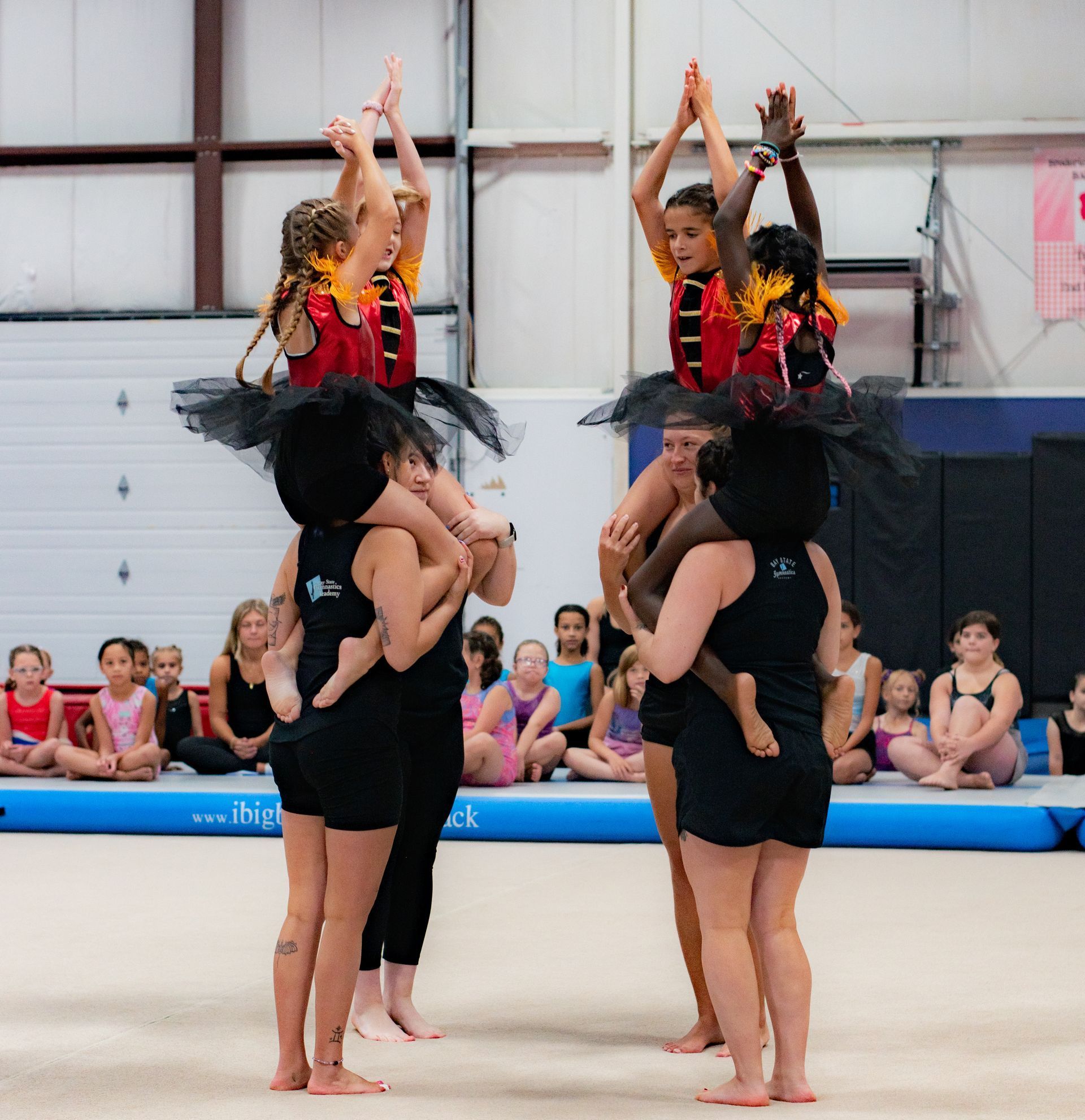 Two cheerleading stunt groups holding teammates aloft with arms raised on an indoor mat.