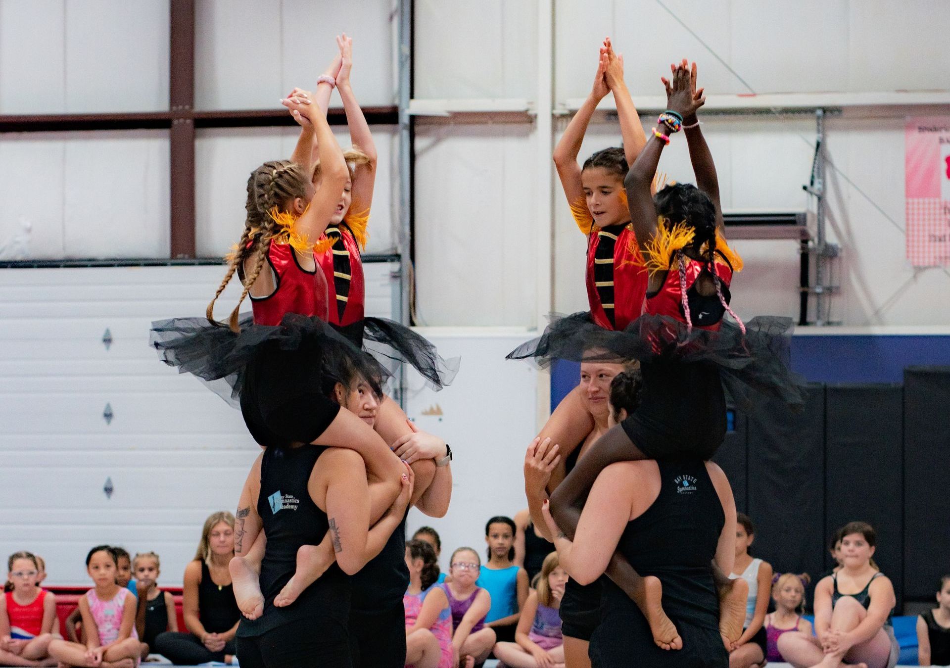 Cheerleaders performing a stunt indoors, with two flyers raised overhead and a seated audience behind them