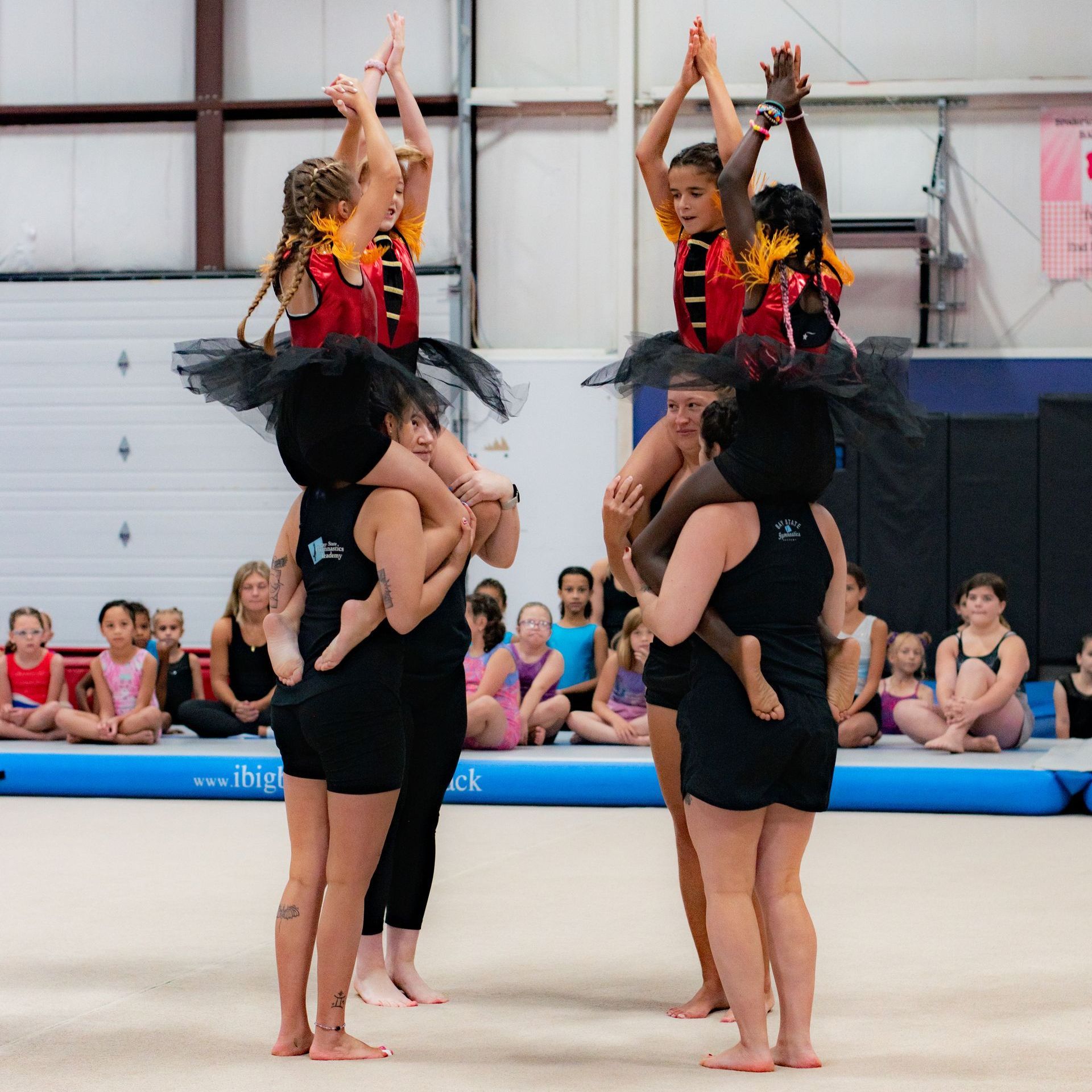 Four gymnasts in black and red perform a stacked acrobatic pose on a gym floor before seated spectators