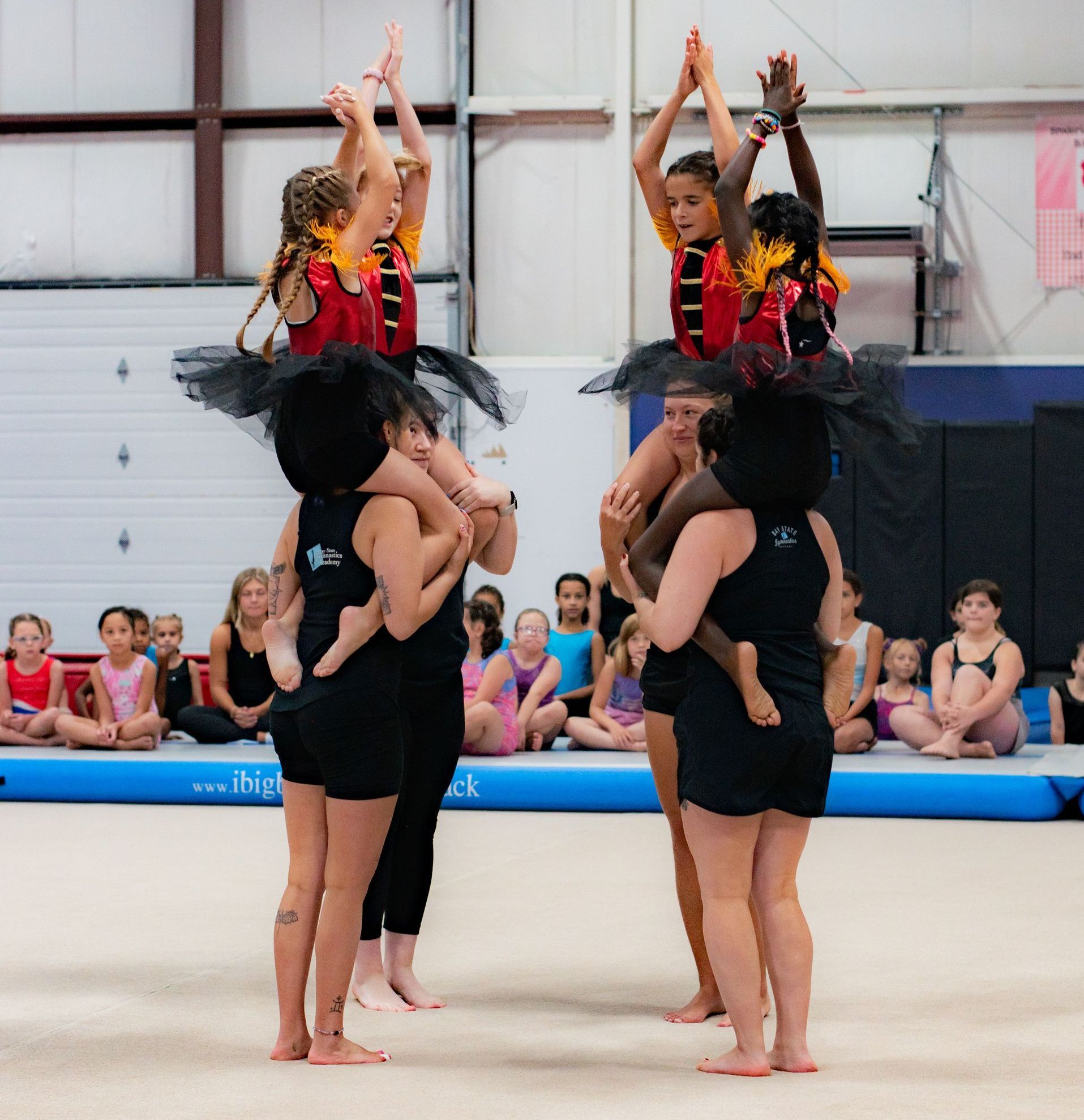Cheerleaders performing a stacked lift routine on a gym floor, with spectators seated in the background