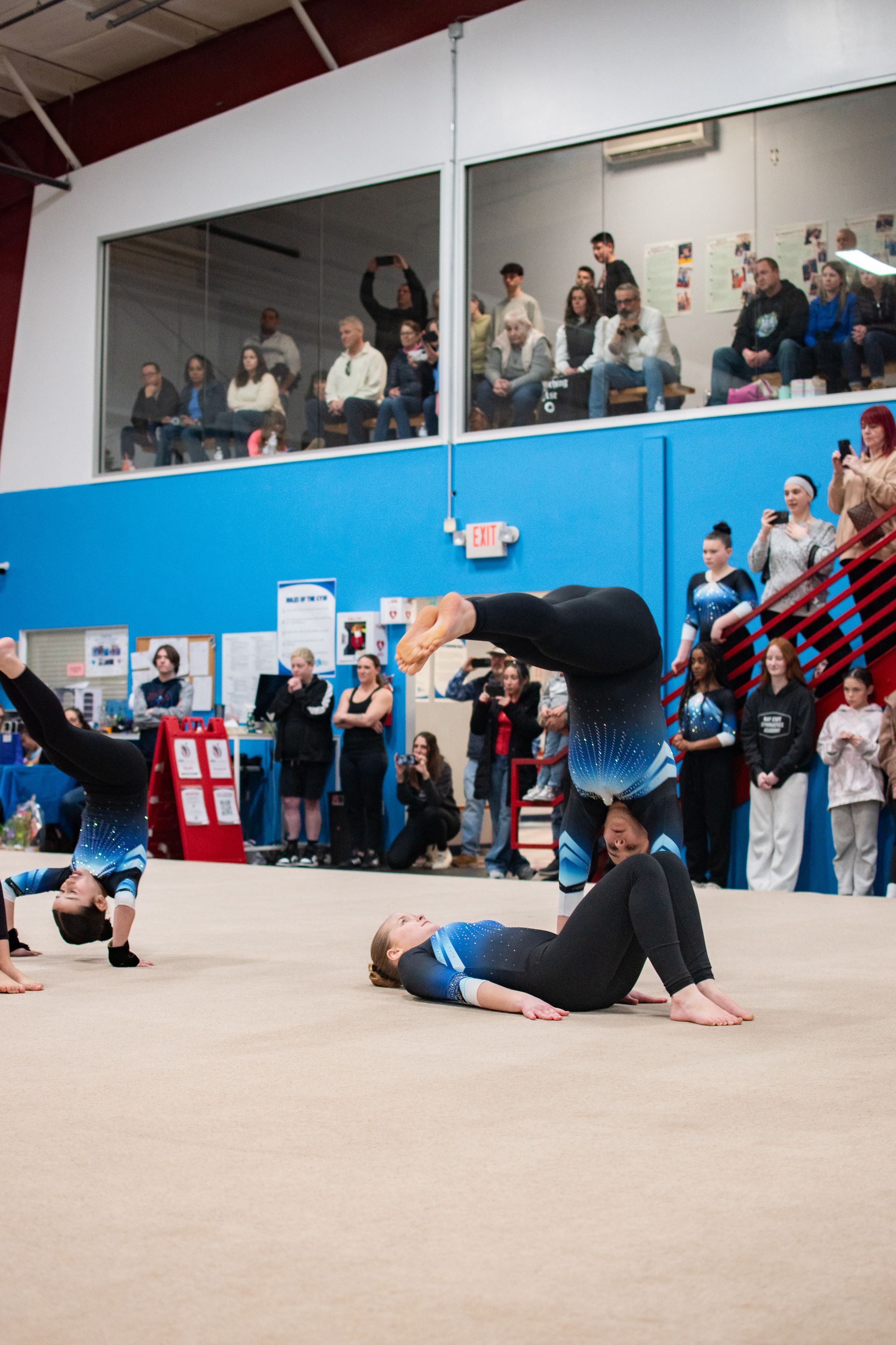 Gymnasts performing a floor routine in a gymnasium, with spectators watching from the stands.