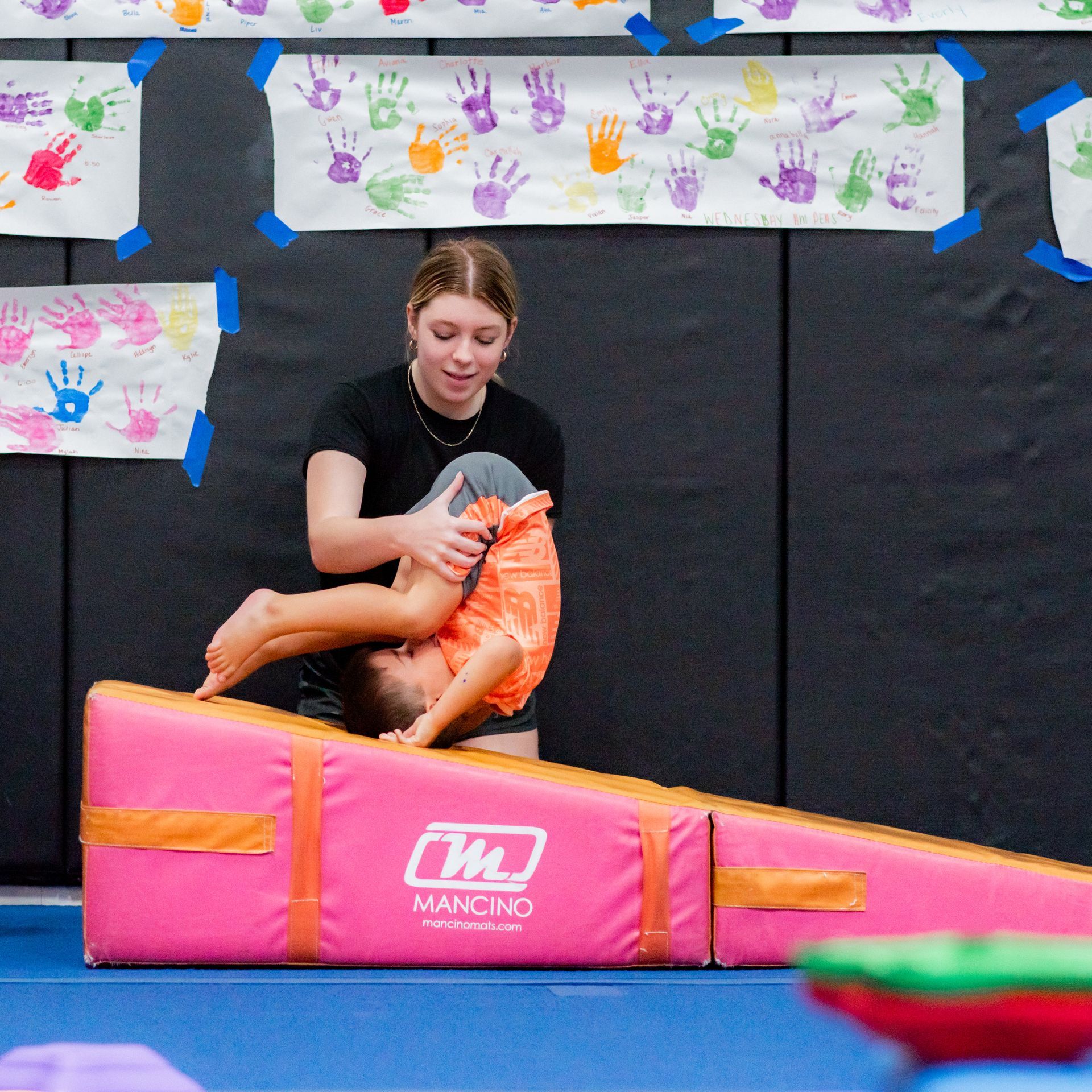 Person in orange outfit crouches on pink padded gym mat, with colorful handprint decorations in the background