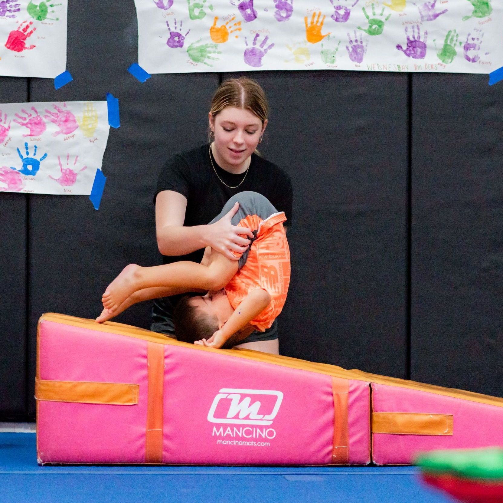 Person sitting on a pink mat, wrapped in an orange blanket, in front of black wall panels and handprint artwork.